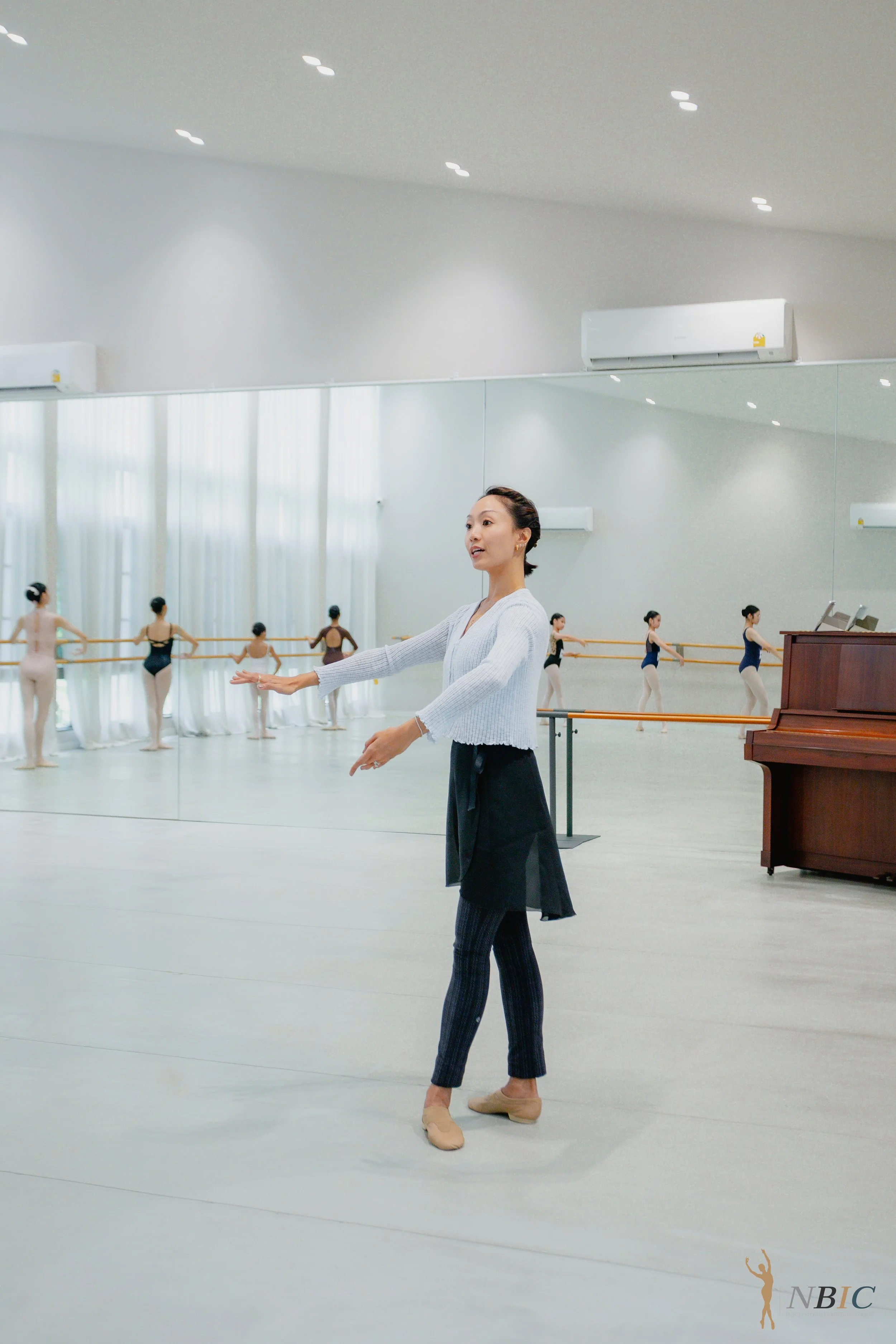 A ballet instructor in a dance studio demonstrating a ballet pose while facing a mirror, with students practicing at the barre in the background.