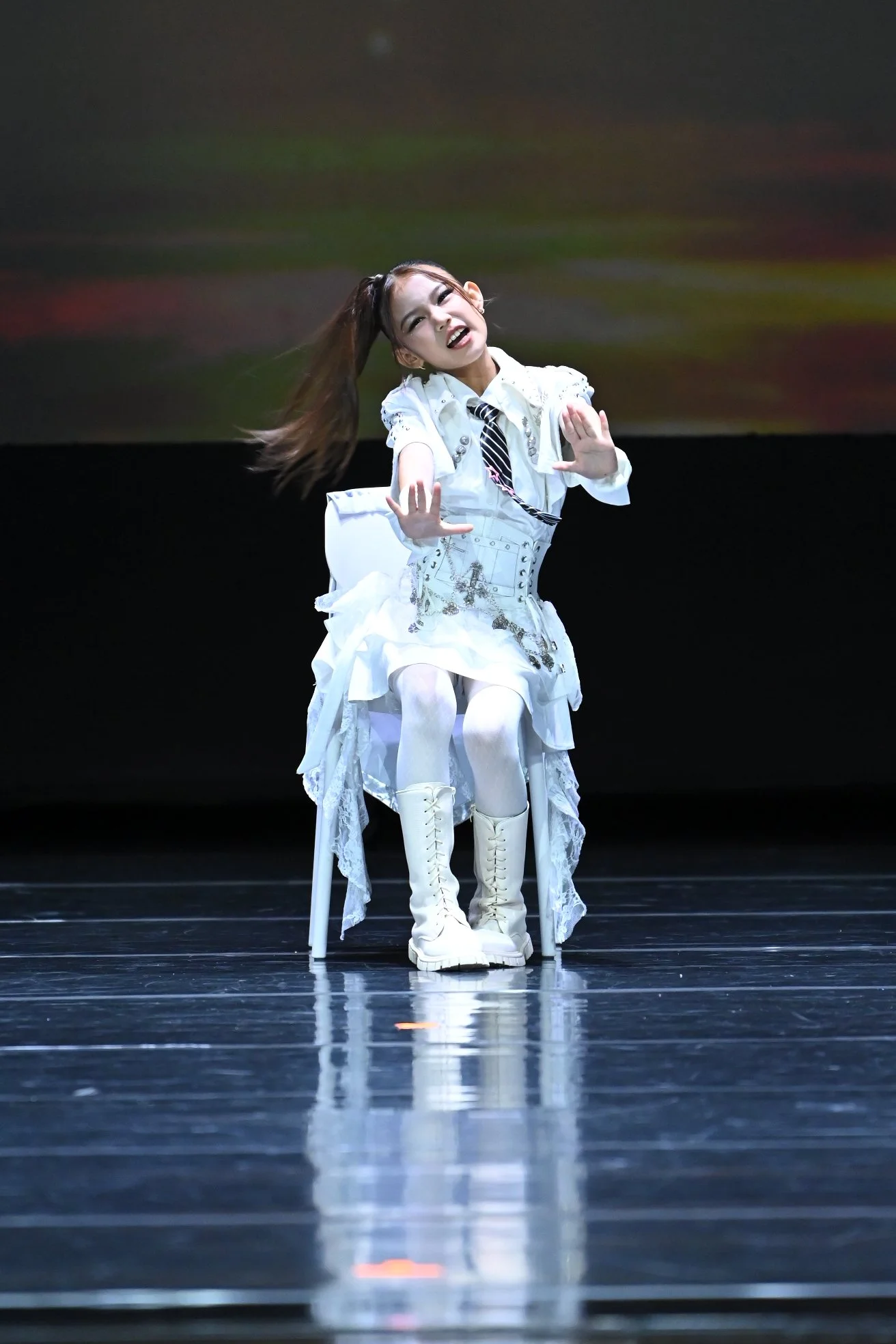 A performer sitting on a white chair on a stage, dressed in a white outfit with decorative details, white thigh-high boots, and a tie, striking a pose with a playful or expressive facial expression and hand gestures.