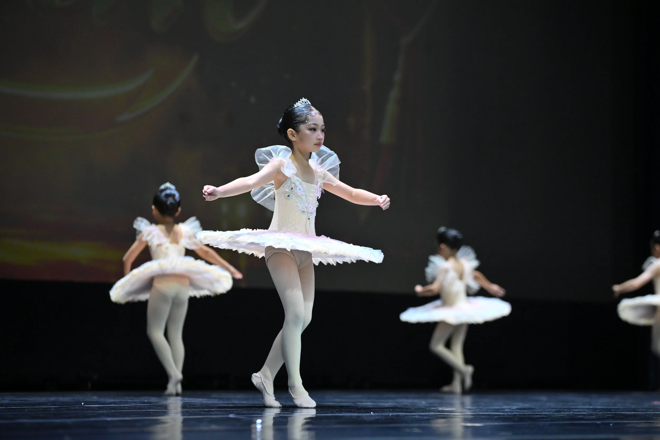 Young ballerina in a cream tutu with silver embellishments, wearing a tiara, practicing ballet on stage with other dancers in the background.