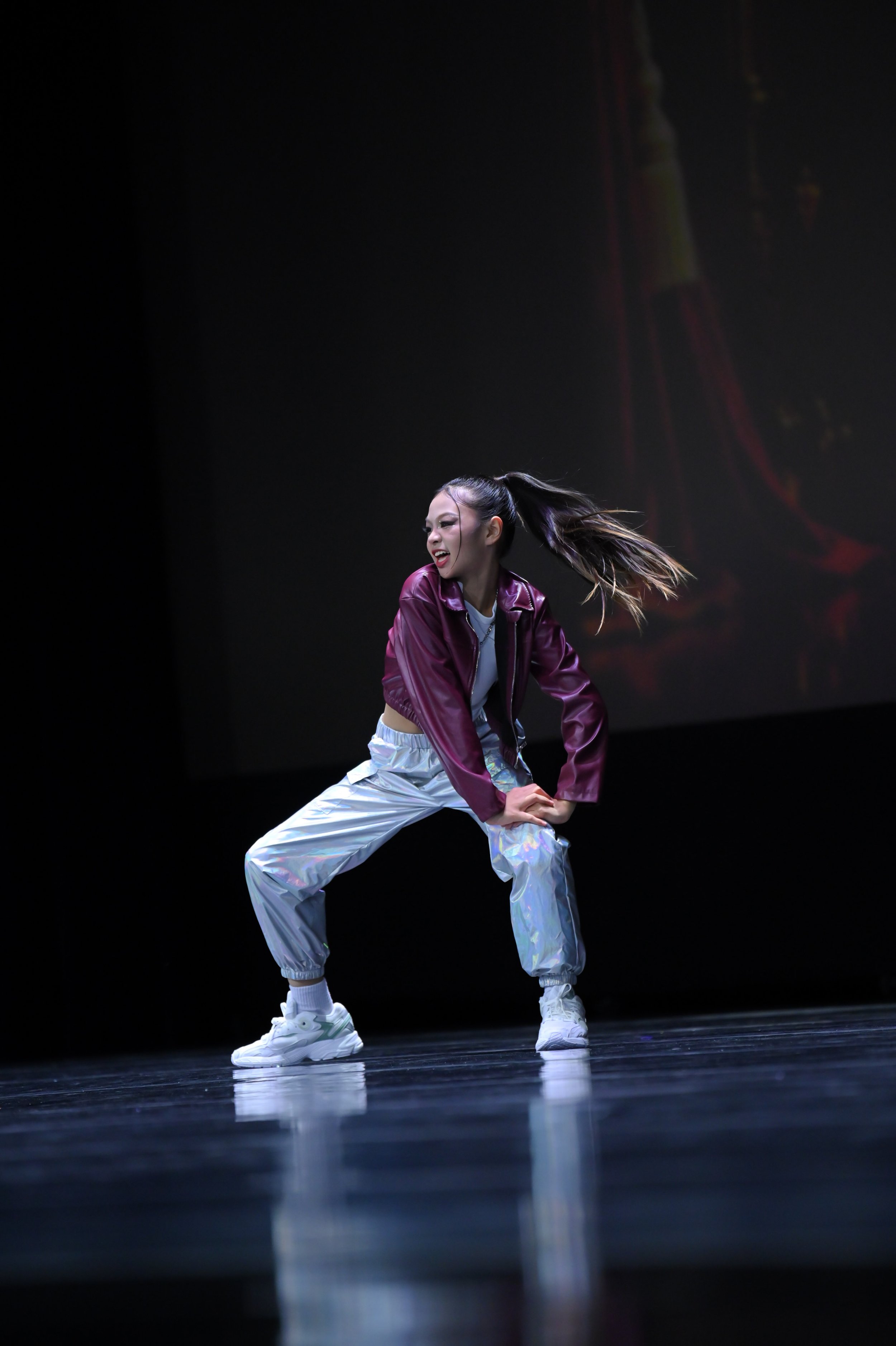 A young female dancer performing on stage, wearing a maroon satin jacket, iridescent pants, and white sneakers, with her hair tied in a high ponytail, against a dark background.