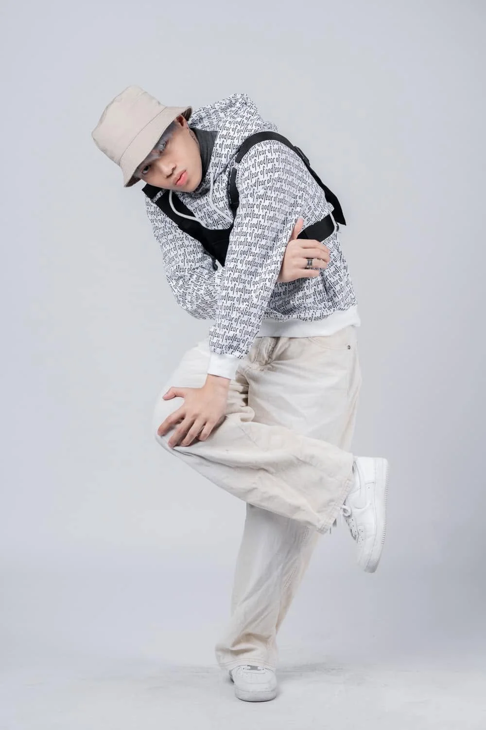 Young man dressed in streetwear with a bucket hat, patterned jacket, and white sneakers, standing against a gray background.