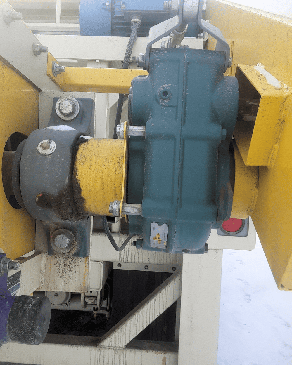 Close-up of a weathered blue gearbox and yellow industrial machinery drive assembly with connected shafts, bolts, and hydraulic components