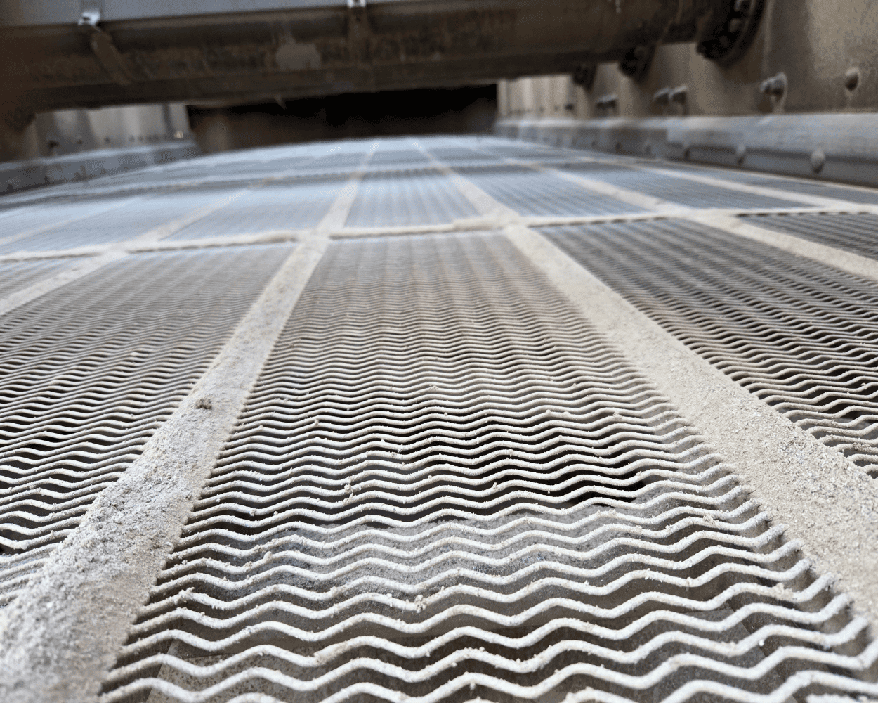 Close-up of a dusty industrial conveyor belt with repeating wavy ridges and metal machinery above