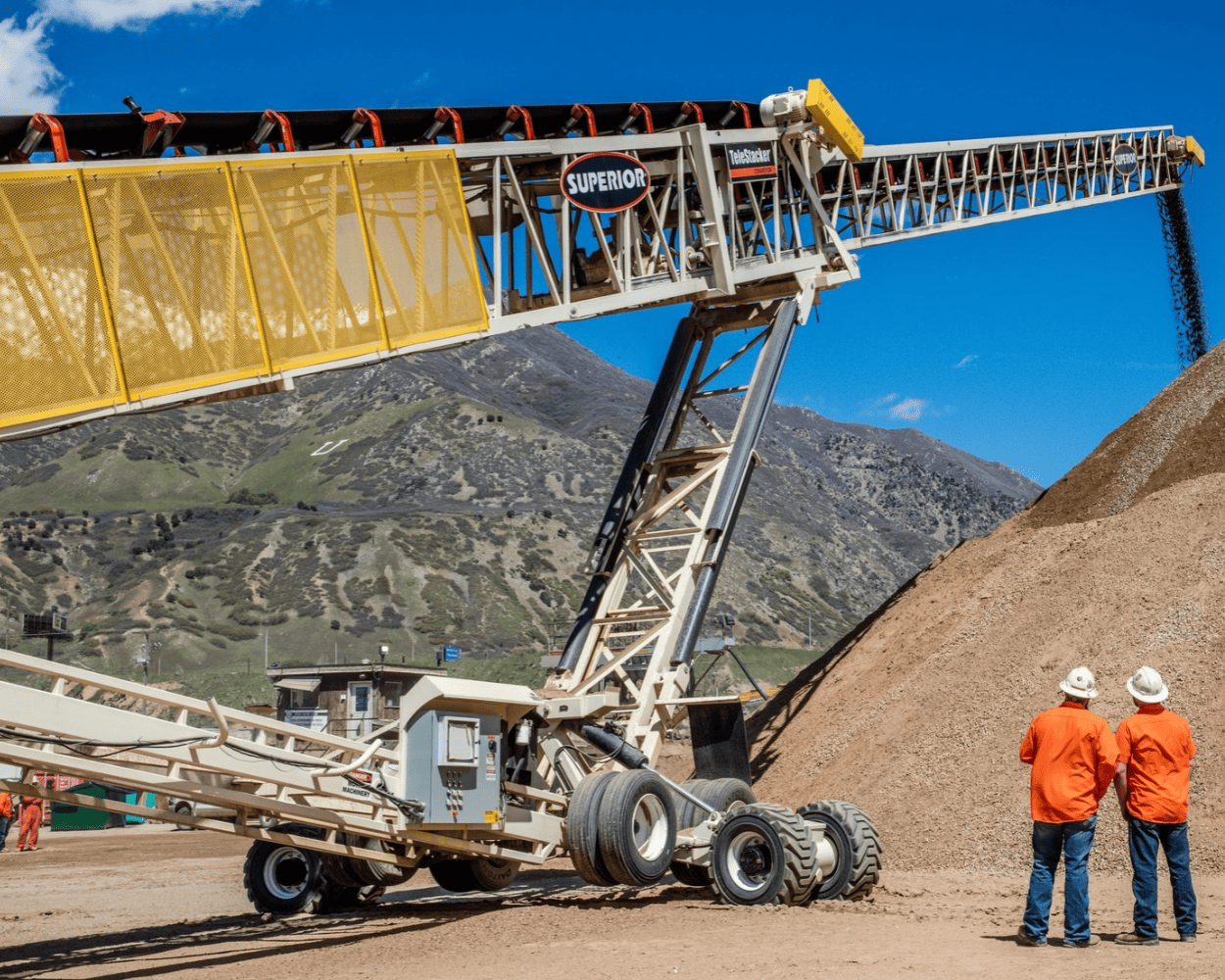 Mobile aggregate conveyor loading a gravel pile at a quarry, with two workers in orange safety jackets and mountain backdrop