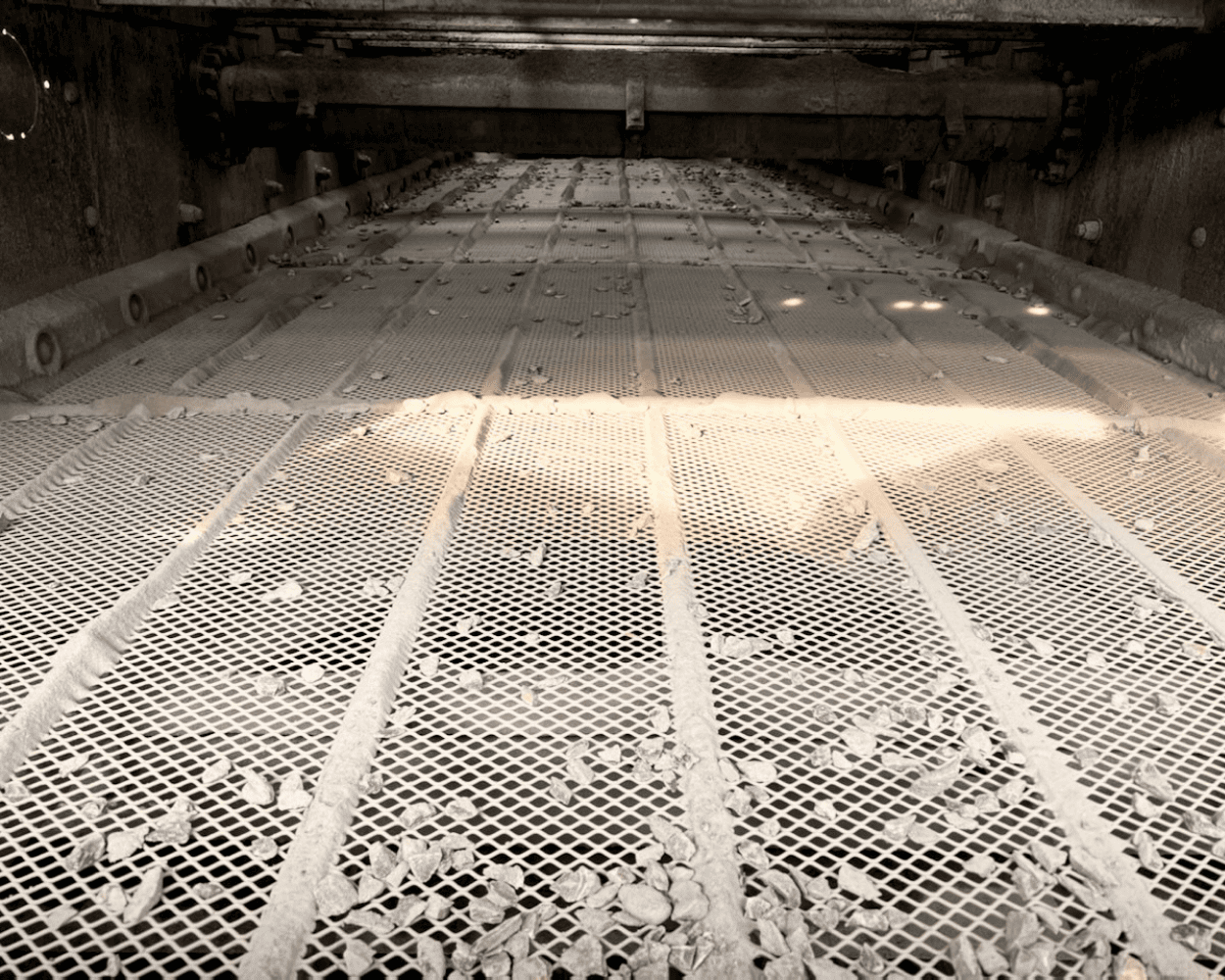 Interior of an industrial processing machine with a perforated metal conveyor belt covered in debris