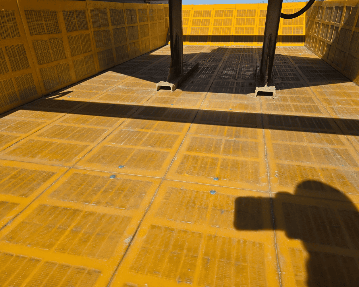 Interior of a yellow industrial metal enclosure with grated panels, support beams, and strong sunlight casting shadows.