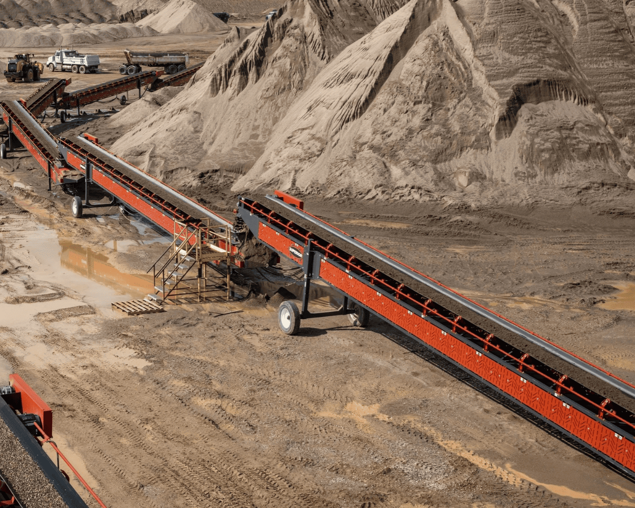 Industrial sand quarry with red conveyor belts, large sand piles, and heavy-duty trucks