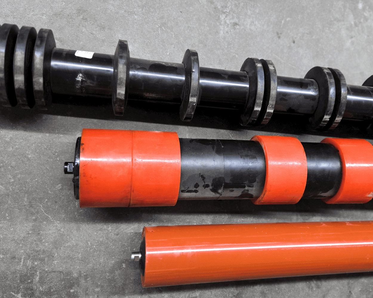 Industrial conveyor rollers and shafts, including black grooved rollers and orange cylindrical rollers, on a concrete floor