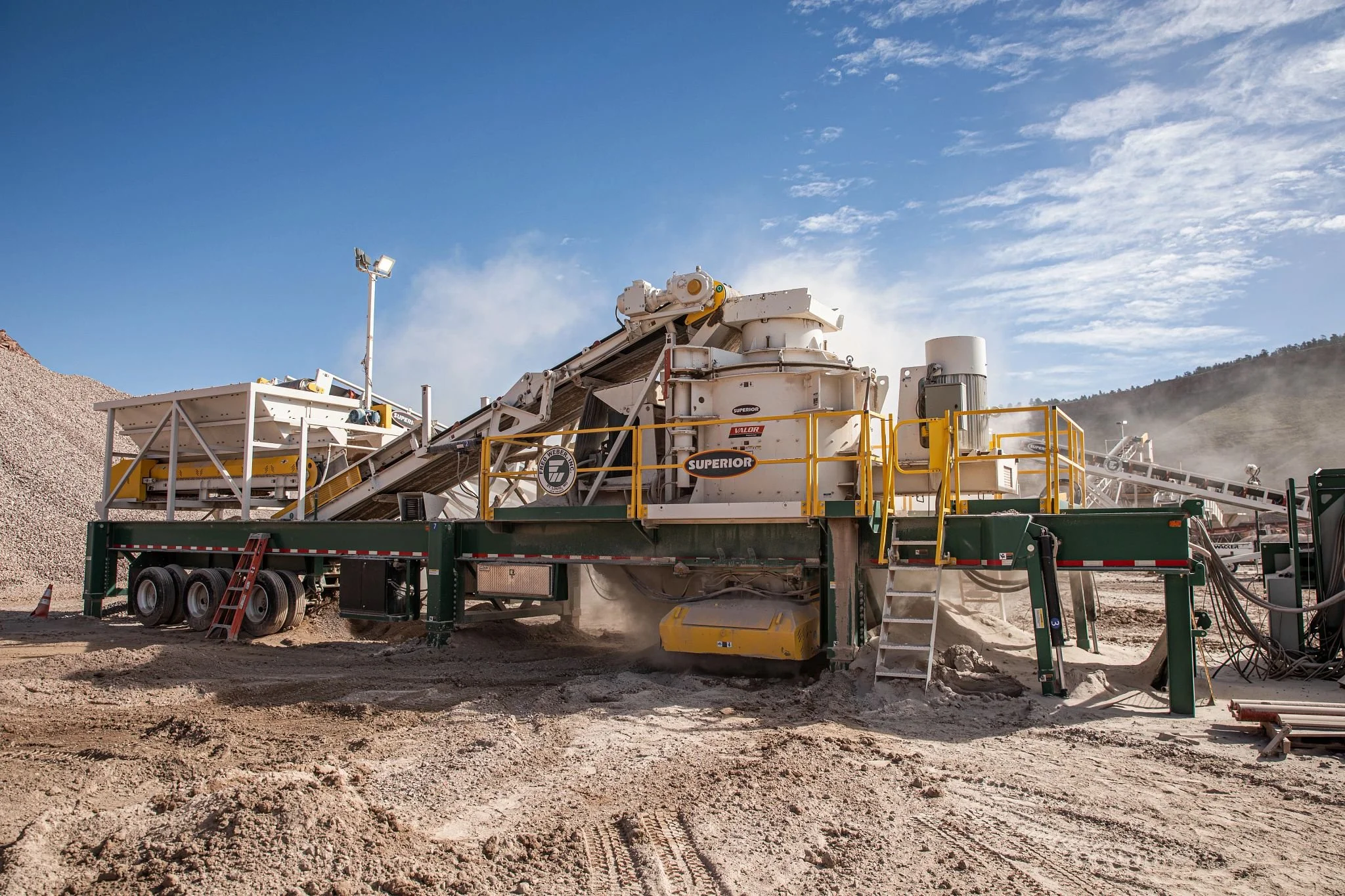 Industrial rock-crushing and screening equipment operating at a dusty quarry under a blue sky