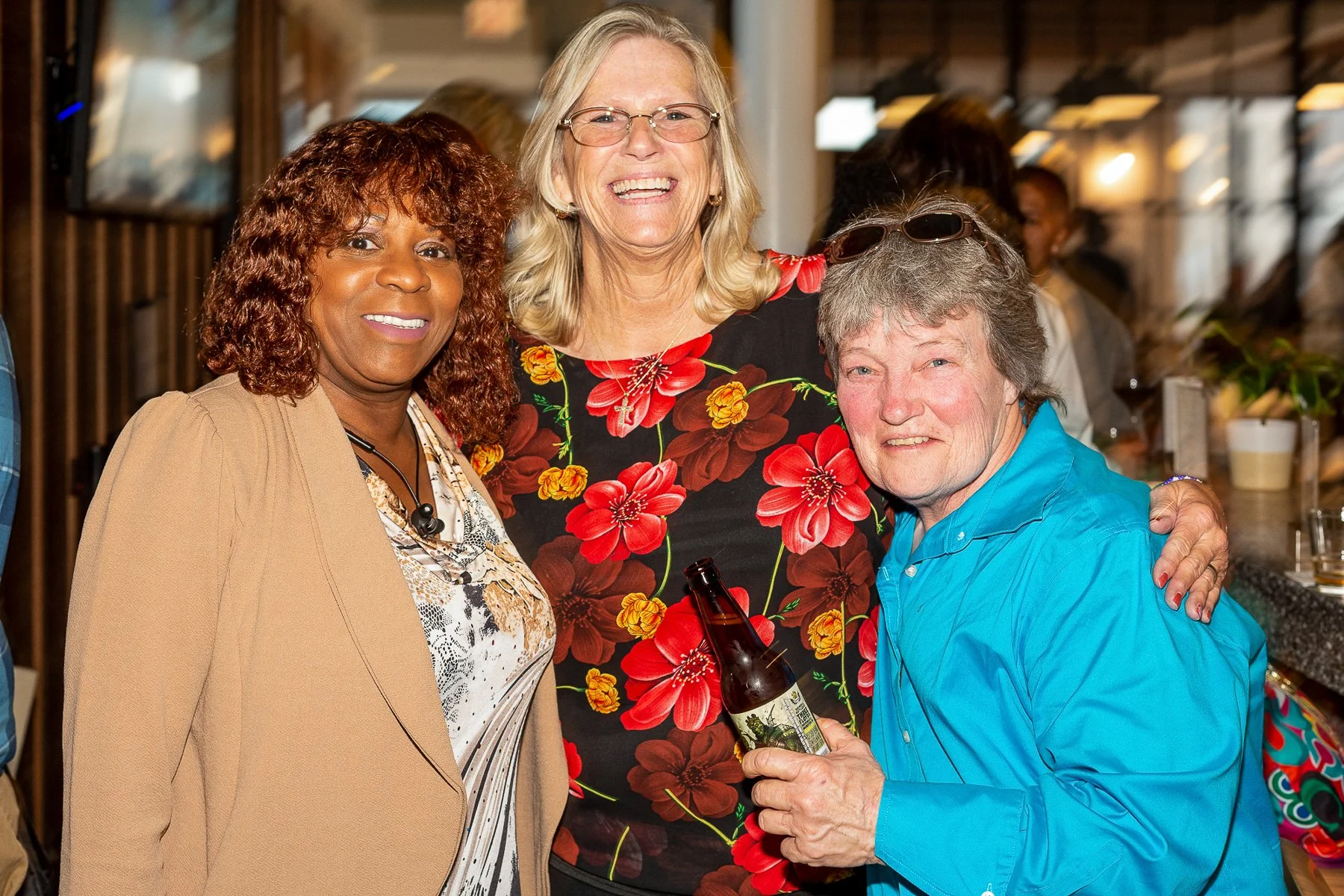 Three people smiling and posing together at a social event. The woman in the middle has blonde hair, glasses, and is wearing a black dress with a large red and yellow floral pattern. The woman on the left has curly brown hair and is wearing a tan bla