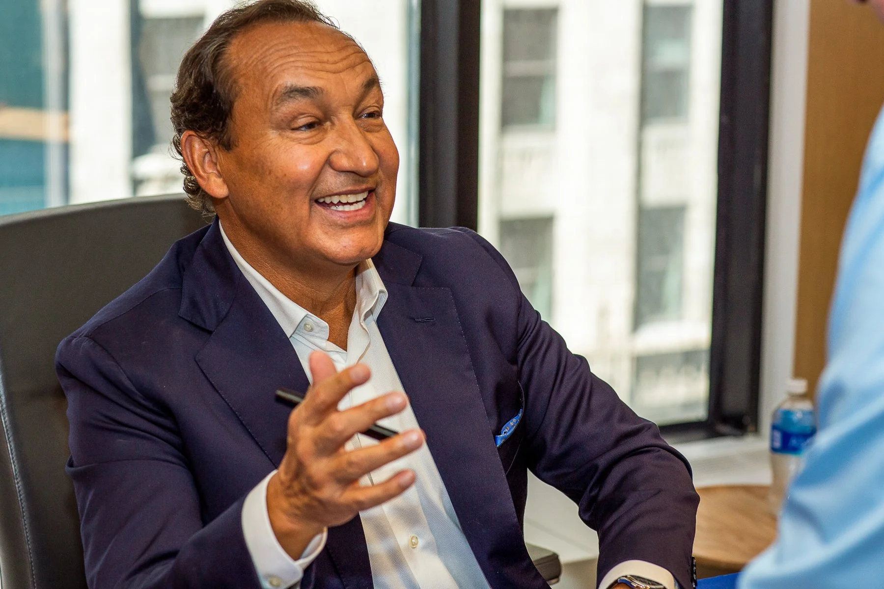 A middle-aged man with light brown skin, wearing a navy blazer and white shirt, is smiling and gesturing with his hand during a conversation in an office setting with large windows in the background.
