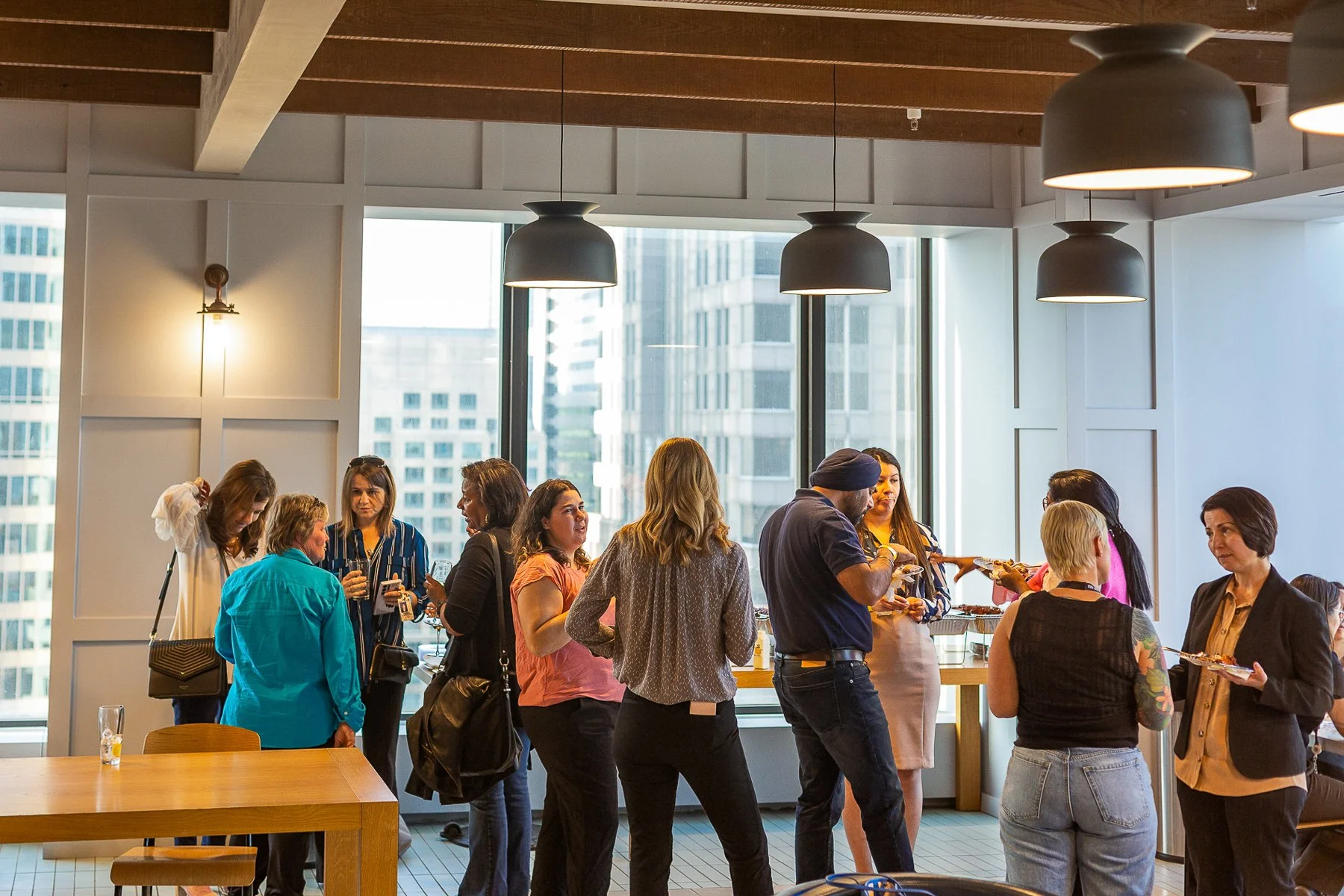 People gathered around a buffet table in a modern high-rise setting, with large windows showing city buildings outside. They are chatting and serving themselves food during a social event or office gathering.