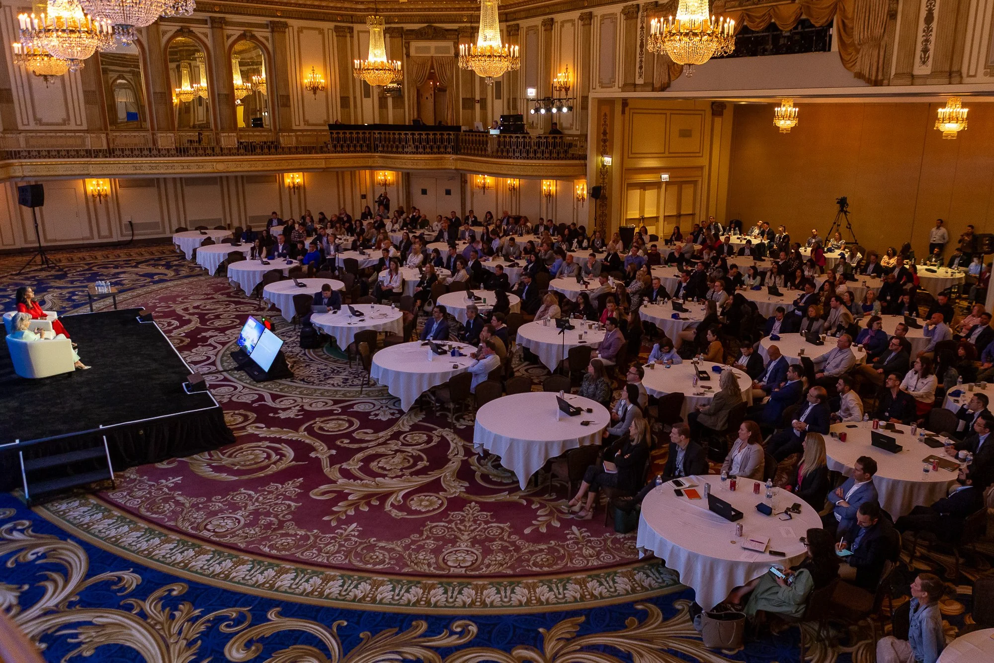 A large conference ballroom filled with round tables and seated attendees listening to a speaker on stage. The room features ornate chandeliers, gold trim, and patterned carpet.©2026 Steven Barston