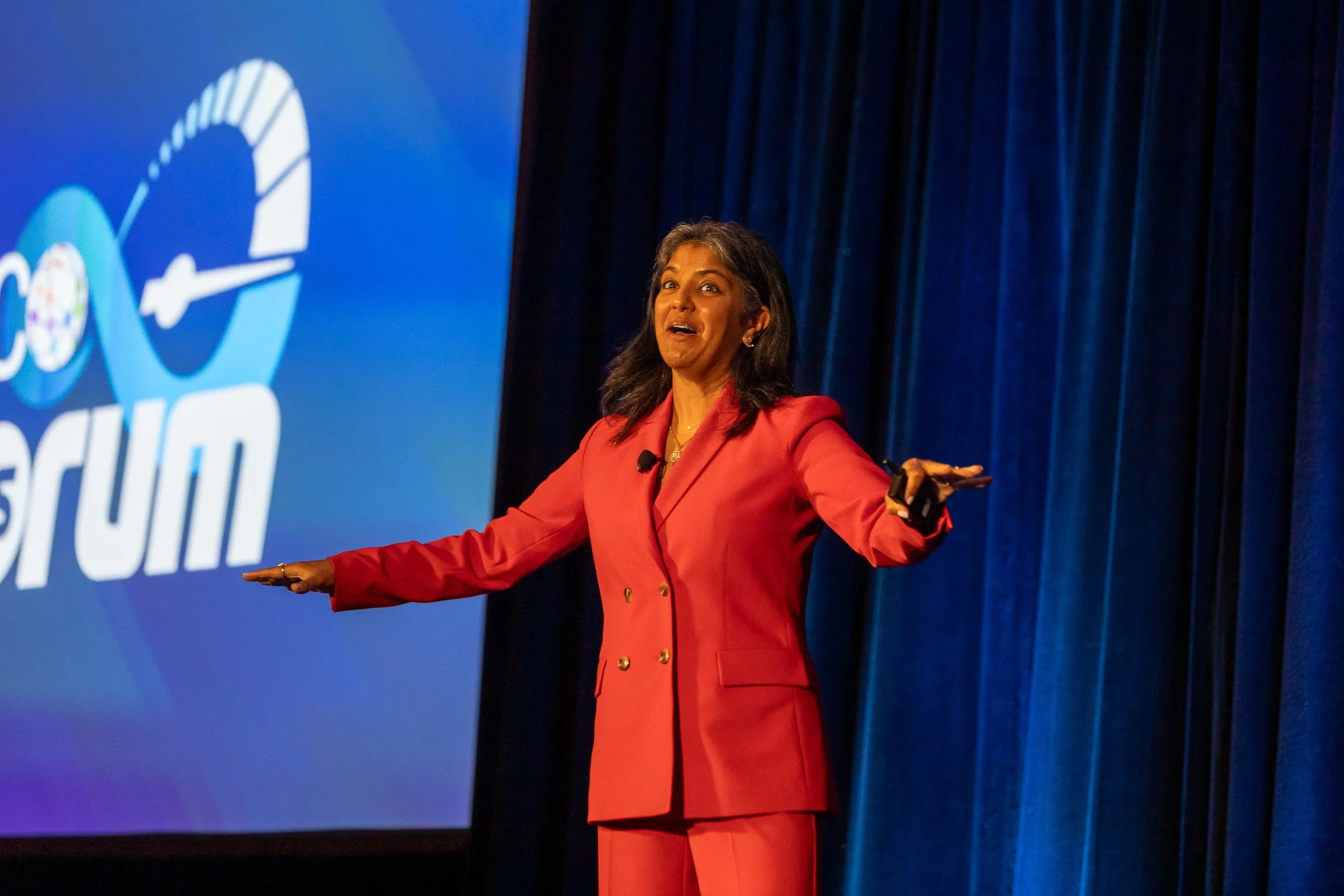 A woman in a red blazer and matching pants is speaking on a stage, gesturing with her hands. Behind her is a large screen displaying a partial logo with a person in front of a blue background with a dark curtain.©2026 Steven Barston
