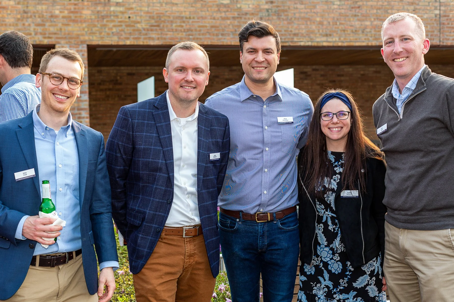 Group of five diverse professionals smiling outdoors at a social or networking event, standing in front of a brick building, with two other people partially visible on the sides.©2026 Steven Barston