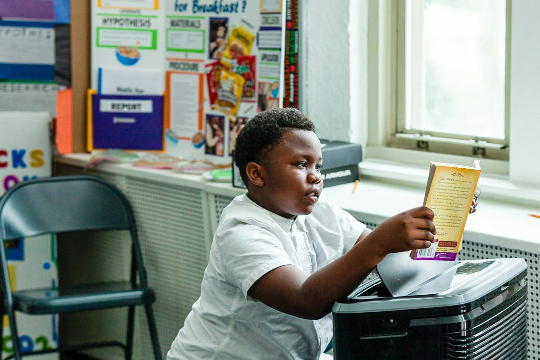A young boy sitting at a desk in a classroom, reading a book, with a bulletin board filled with notices and colorful papers behind him, and an open window letting in natural light.