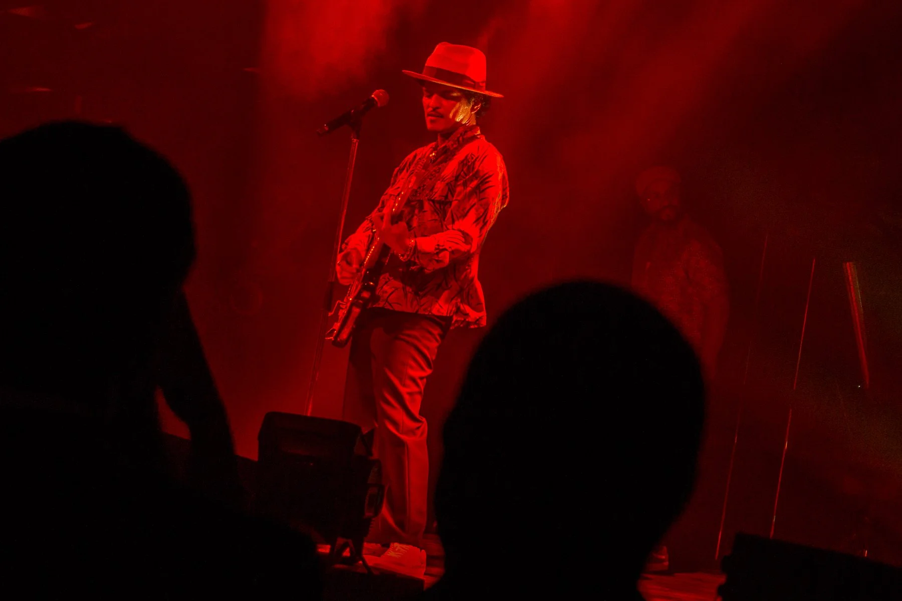 Musician performing on stage with red lighting, wearing a wide-brimmed hat, patterned jacket, and holding a guitar, with silhouettes of audience members visible in the foreground.