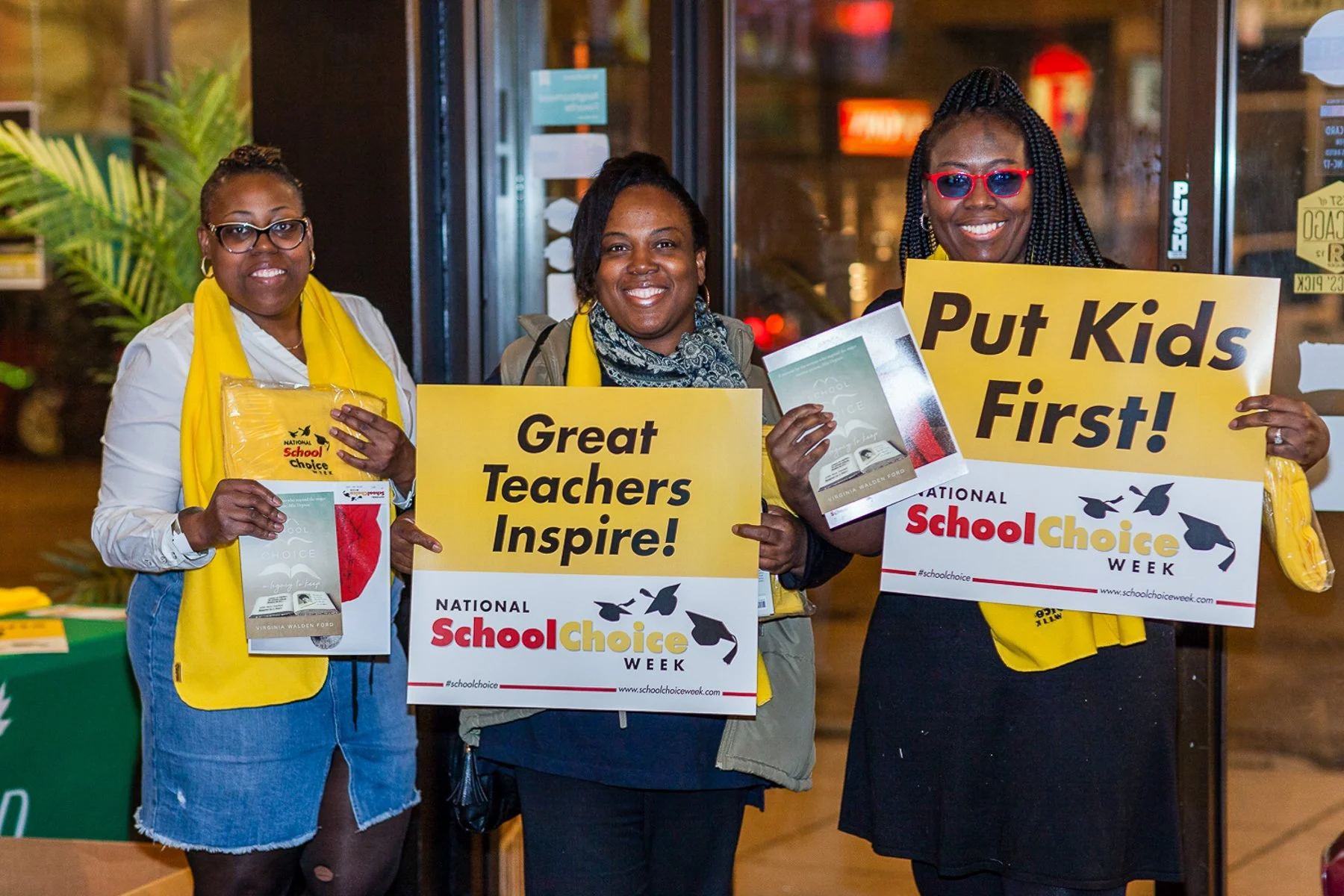 Three women smiling and holding signs for National School Choice Week, wearing yellow scarves. One woman holds a pamphlet, and another holds a sign that says, 'Put Kids First!' The background shows a glass door and some greenery.