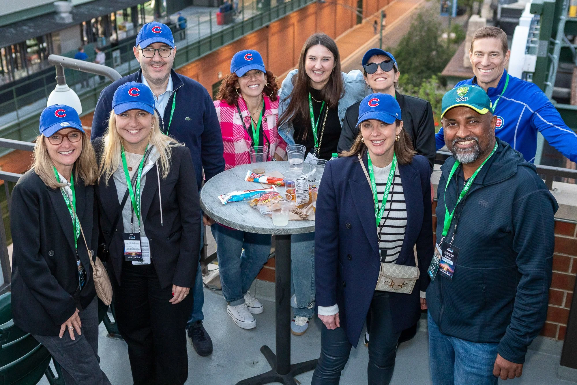 Group of ten people, five women and five men, standing around a high table outdoors, smiling for the camera. They are wearing Chicago Cubs baseball caps and various casual and professional clothing. Snack food and drinks are on the table.©2026 Steven