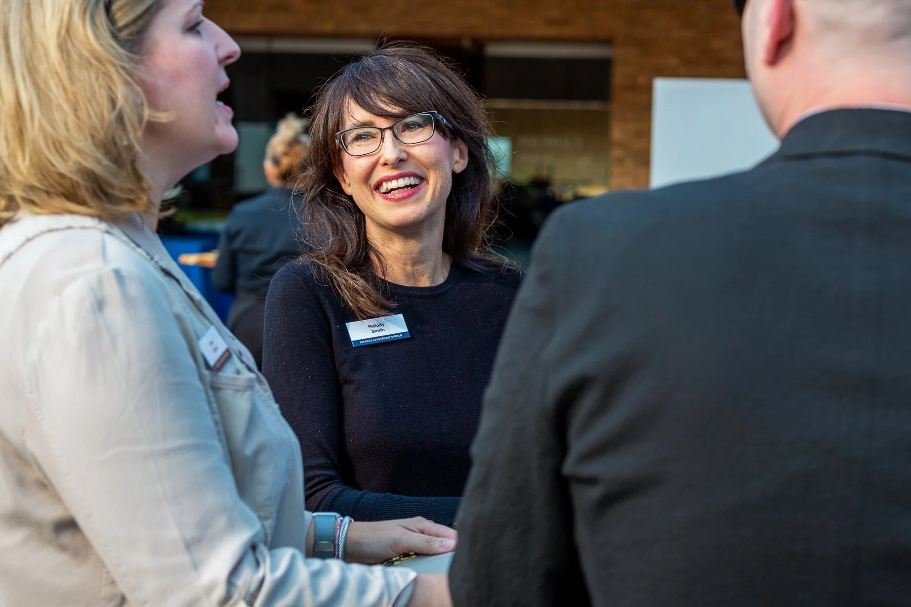 A group of professionally dressed people engaged in a conversation at a networking event, with a woman in the center smiling and wearing glasses and a name tag.©2026 Steven Barston