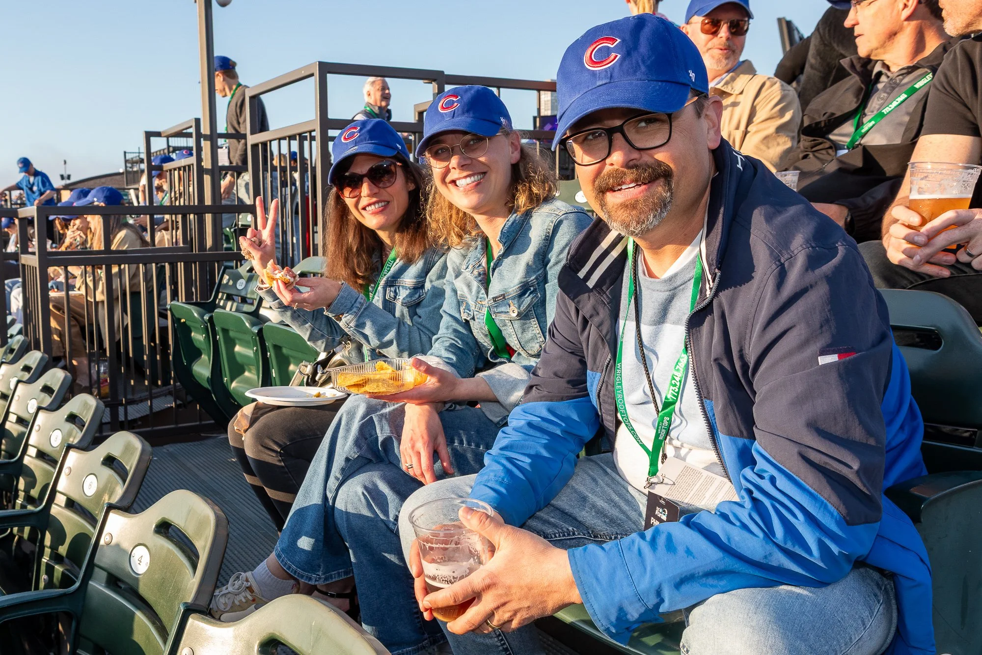©2026 Steven Barston Group of baseball fans sitting in stadium seats, wearing Chicago Cubs caps, eating snacks, and enjoying the game on a sunny day.