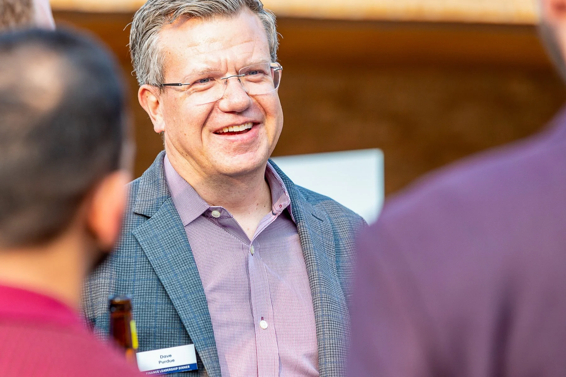 Smiling man wearing glasses and a checkered blazer speaking to people at a professional event.©2026 Steven Barston