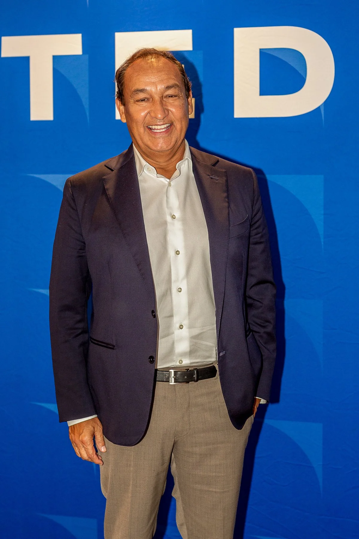 Man in a dark suit jacket, light-colored pants, white shirt, smiling and standing in front of a blue background with large white letters.©2026 Steven Barston