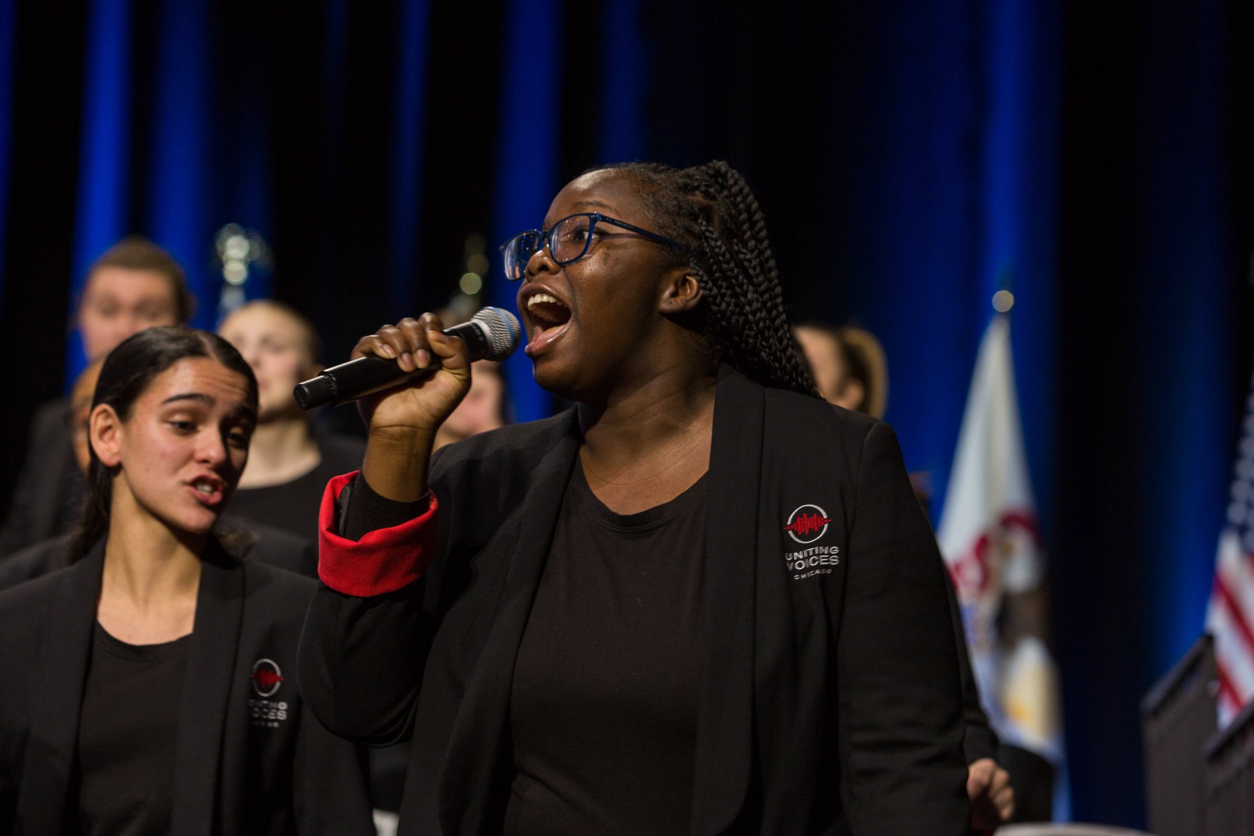 A woman with glasses and braids speaking into a microphone at a rally or event, wearing a black jacket with a logo, surrounded by other women, with American flags and blue curtains in the background.