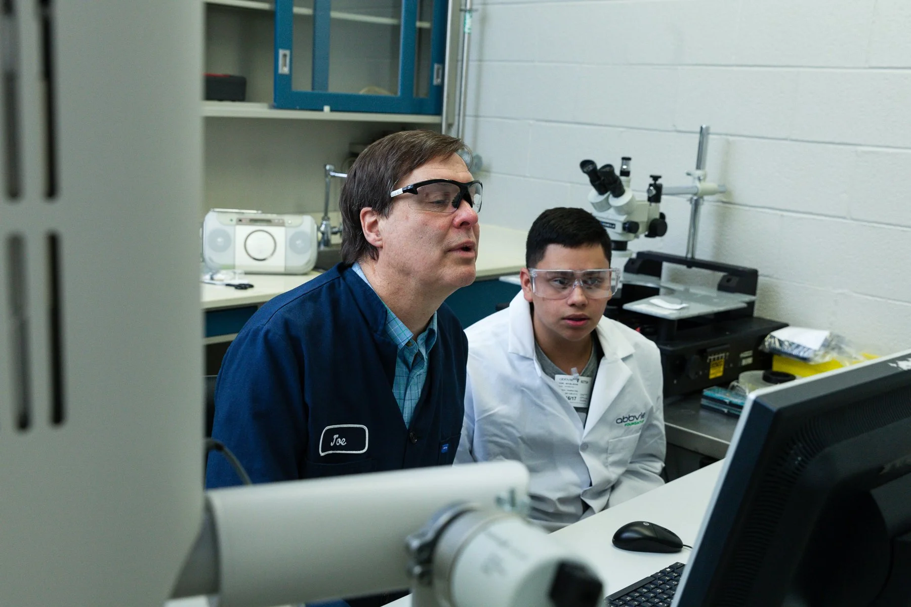 Two scientists, a man and a boy, working together in a laboratory, both wearing safety glasses and focused on a computer screen.