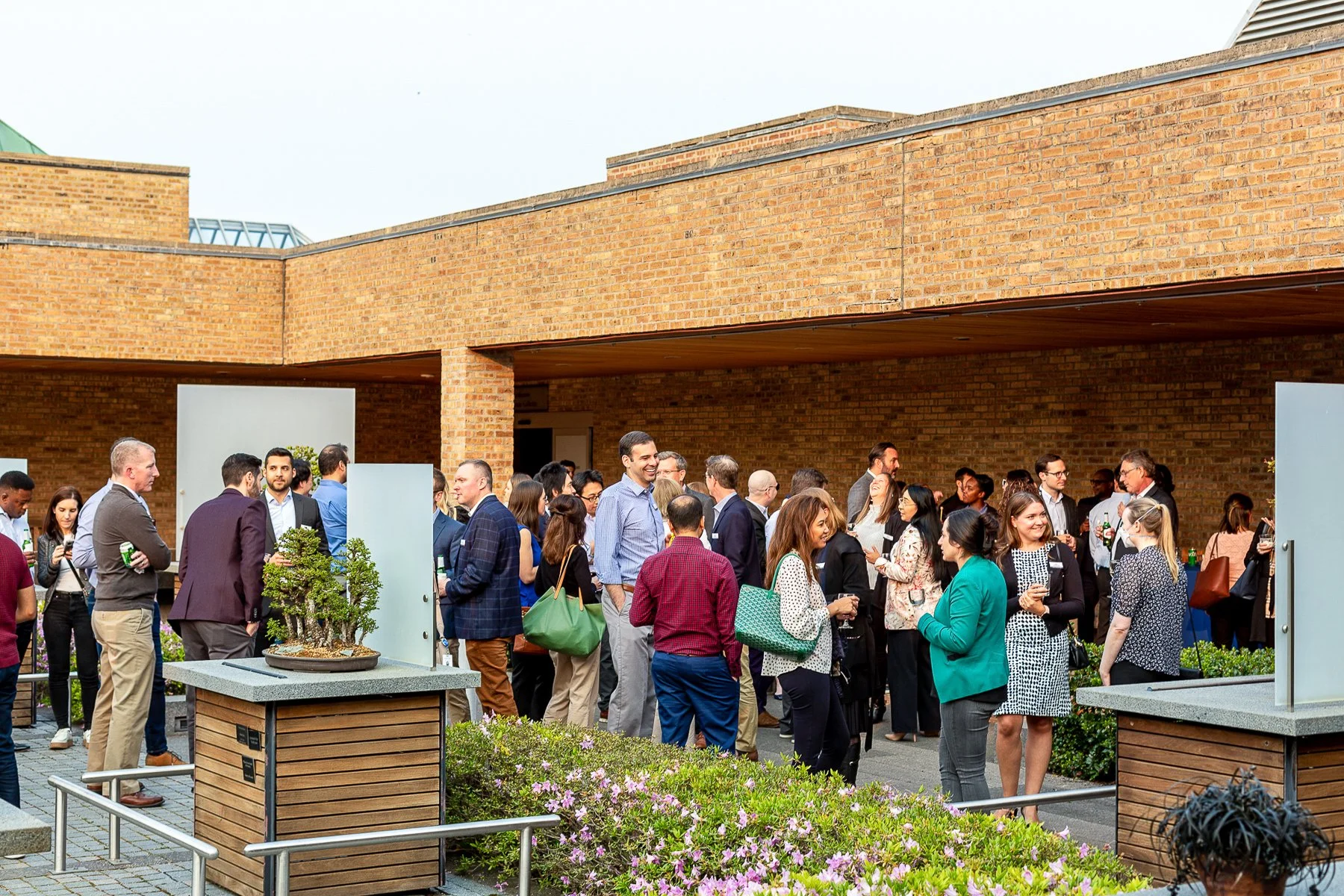 A diverse group of people at an outdoor social event, standing and talking in a patio area with brick walls and some plants.©2026 Steven Barston