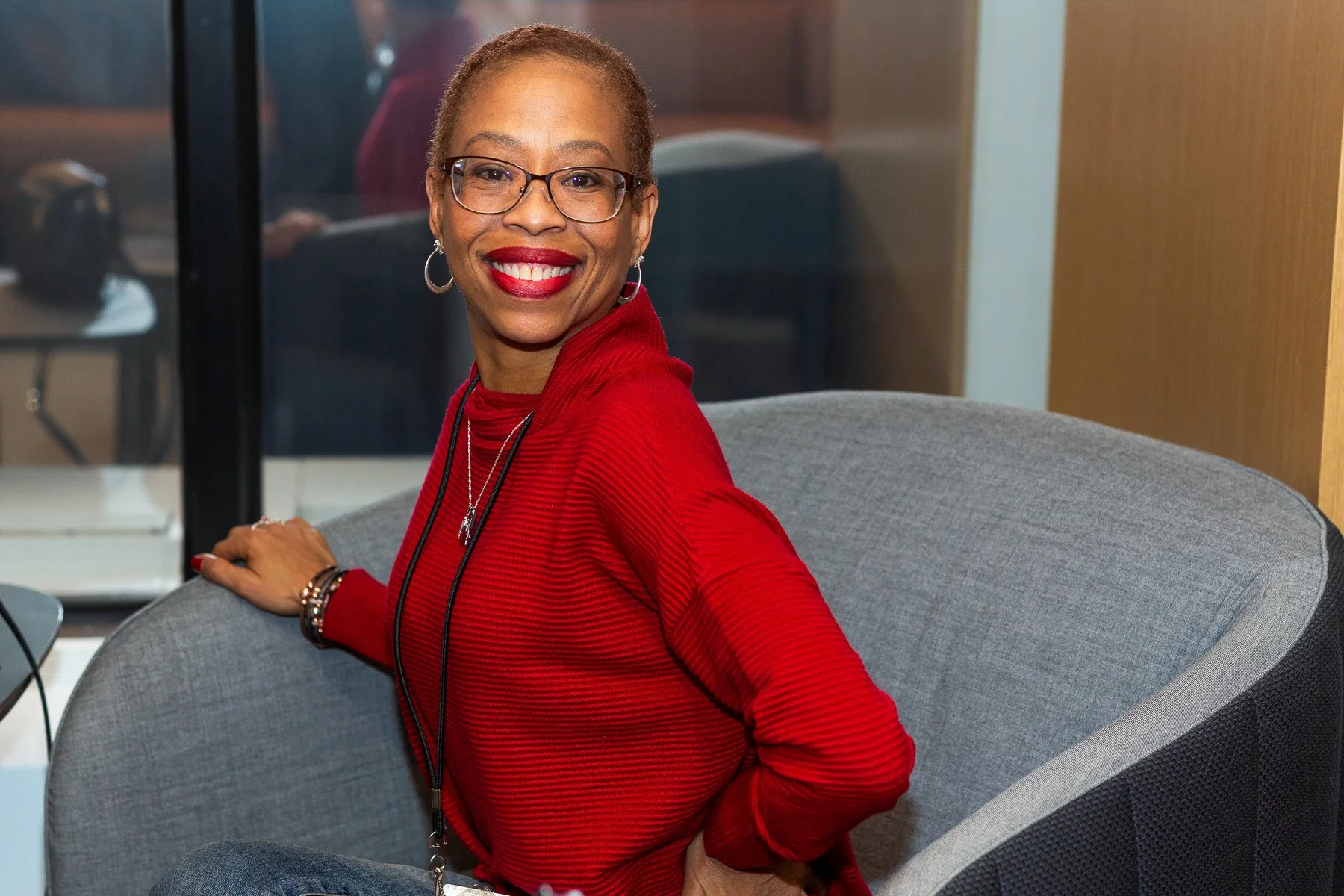 A smiling African American woman with glasses, red lipstick, and short hair, wearing a red striped top and jewelry, sitting on a gray couch in a modern office or lounge area.