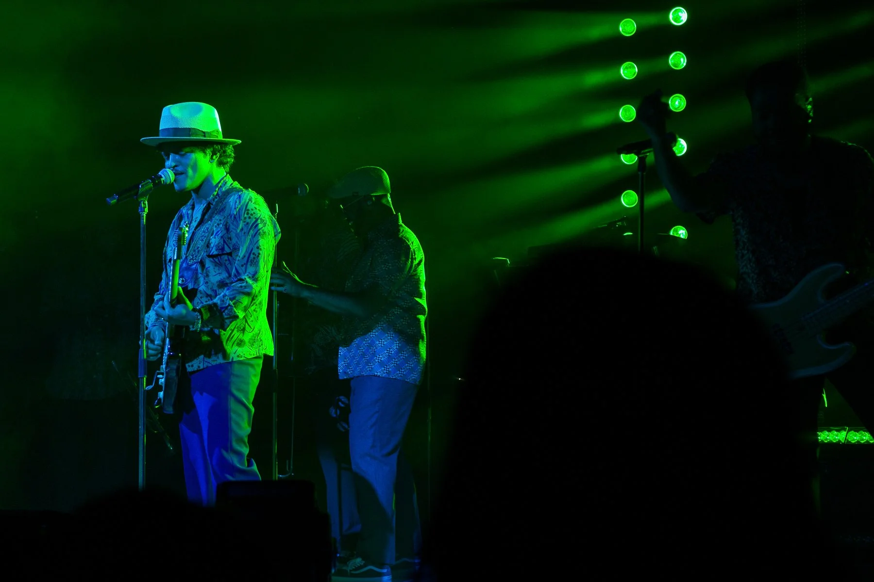 Three musicians performing on stage with green lighting, one playing guitar and singing into a microphone while wearing a hat, another playing bass and wearing glasses, and the third holding a microphone.
