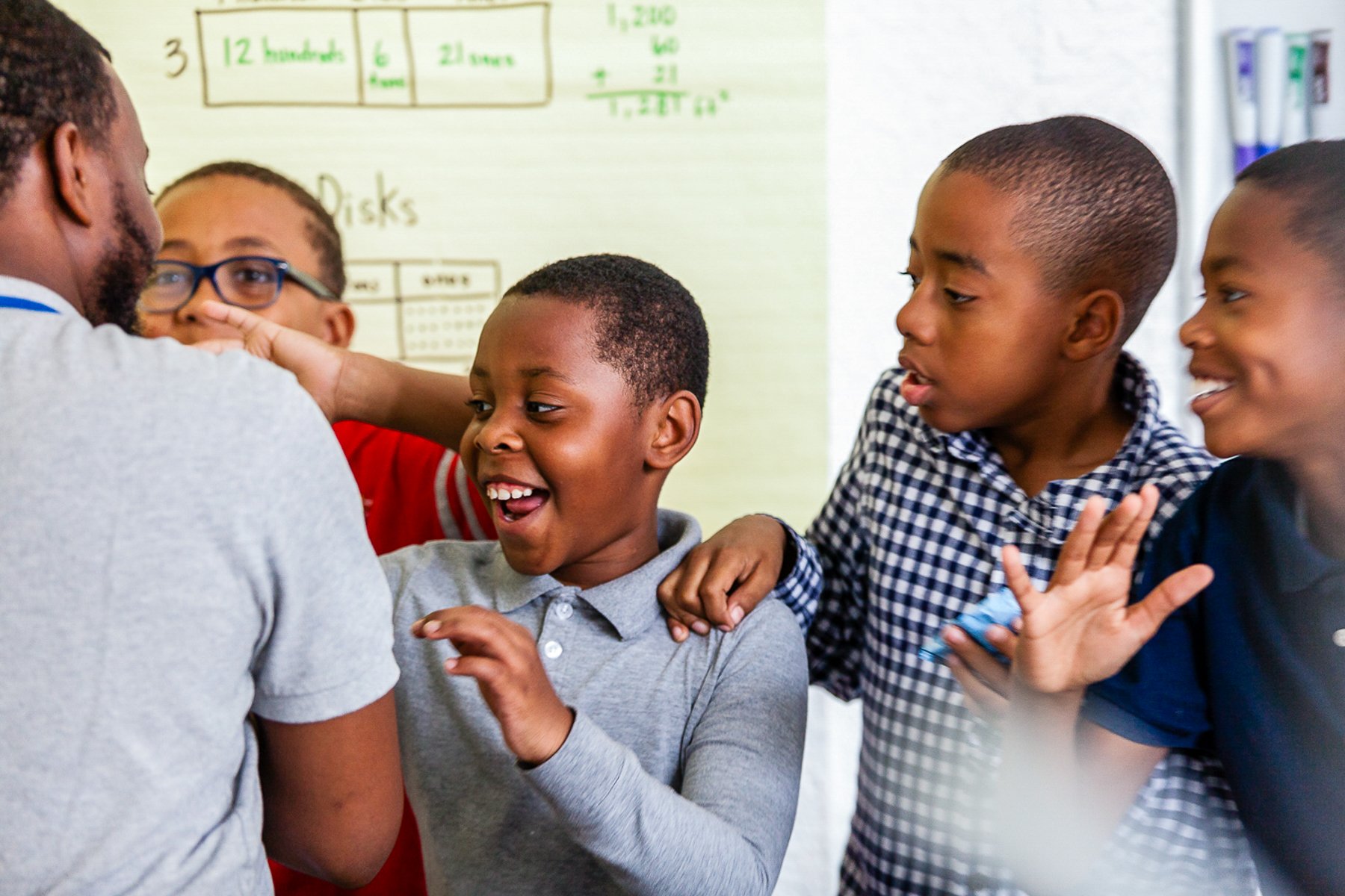 A group of children and an adult smiling and interacting in a classroom, with a whiteboard and educational materials in the background.