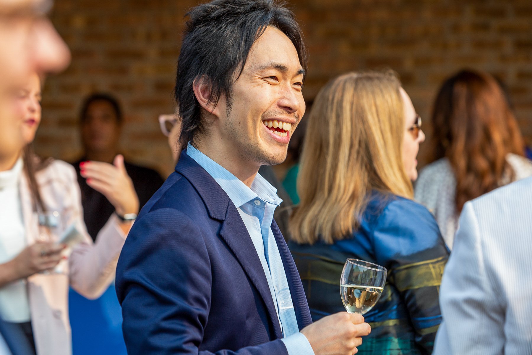 Man smiling and holding a glass of white wine at a social event, surrounded by other people in a lively indoor setting.©2026 Steven Barston