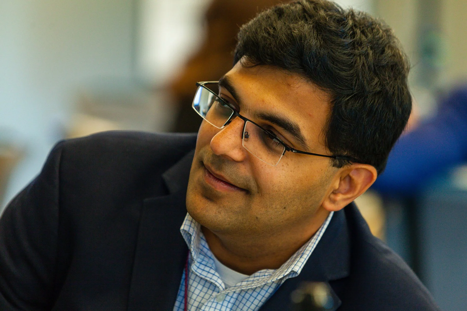 Close-up of a young man with dark hair and glasses, dressed in a dark blazer and a checkered shirt, smiling and looking to his right in an indoor setting.