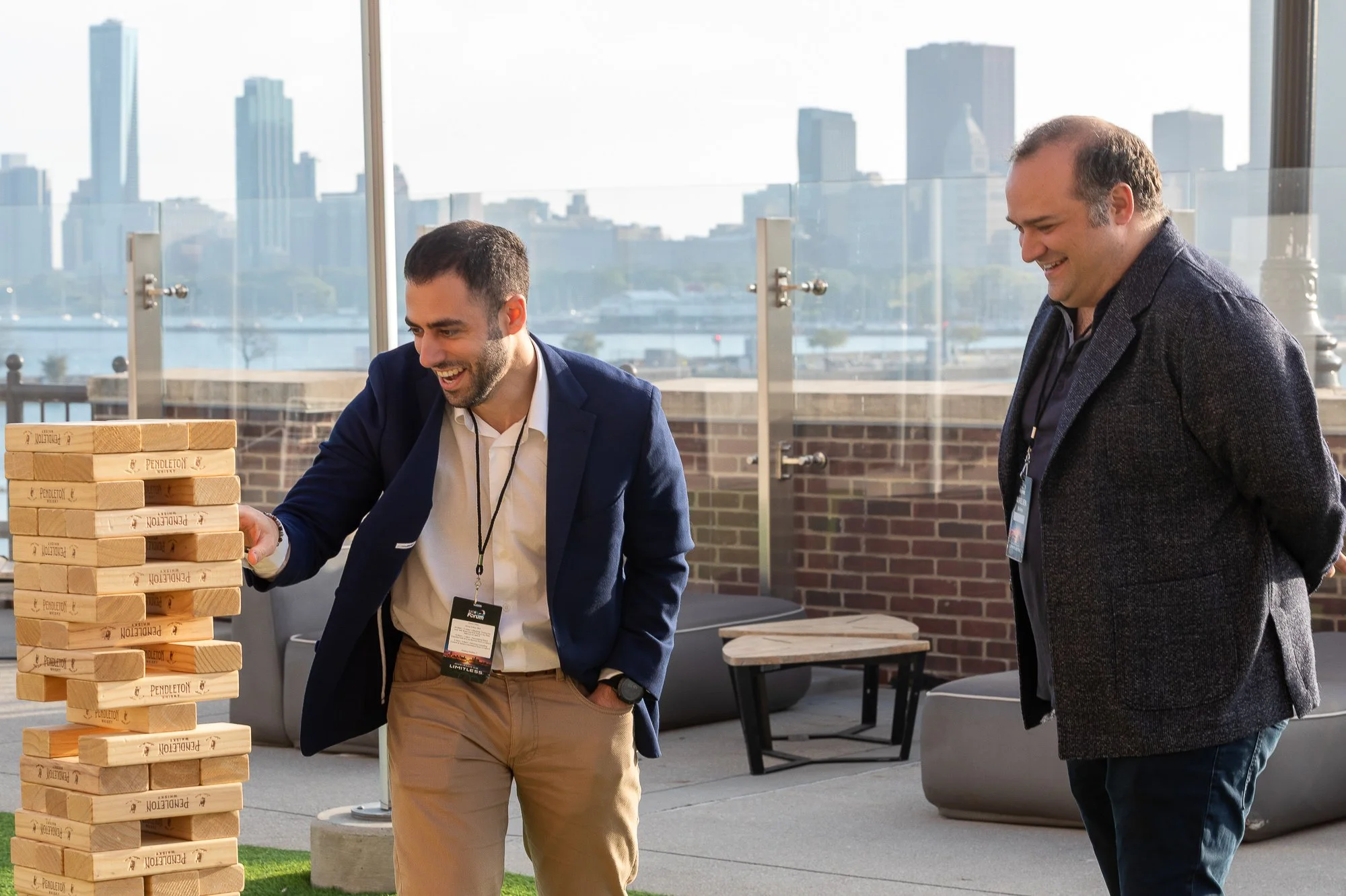 Two men play a giant outdoor game of Jenga on a rooftop with a city skyline in the background. They are laughing and enjoying themselves. ©2026 Steven Barston