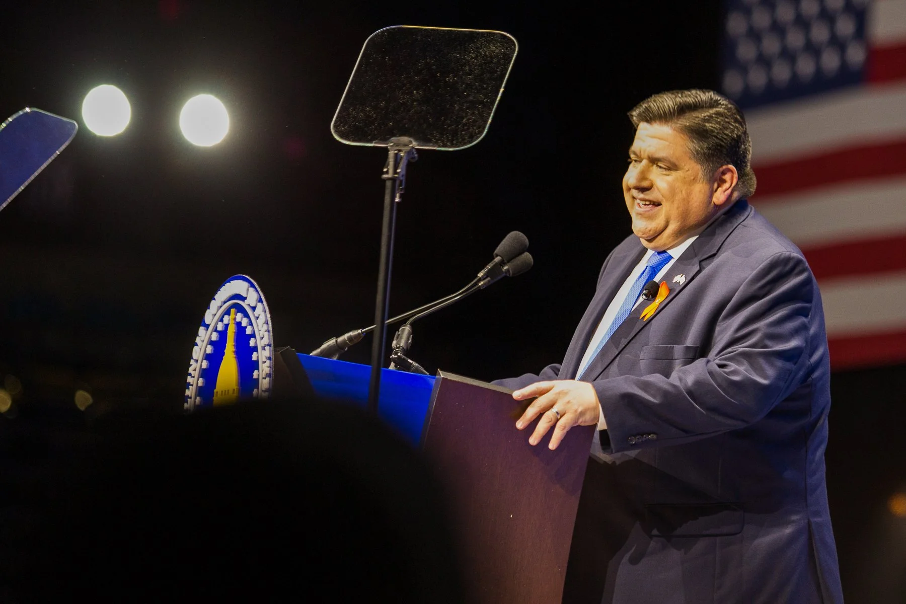 A man in a dark blue suit and a blue tie speaking at a podium with a microphone, with an American flag in the background.