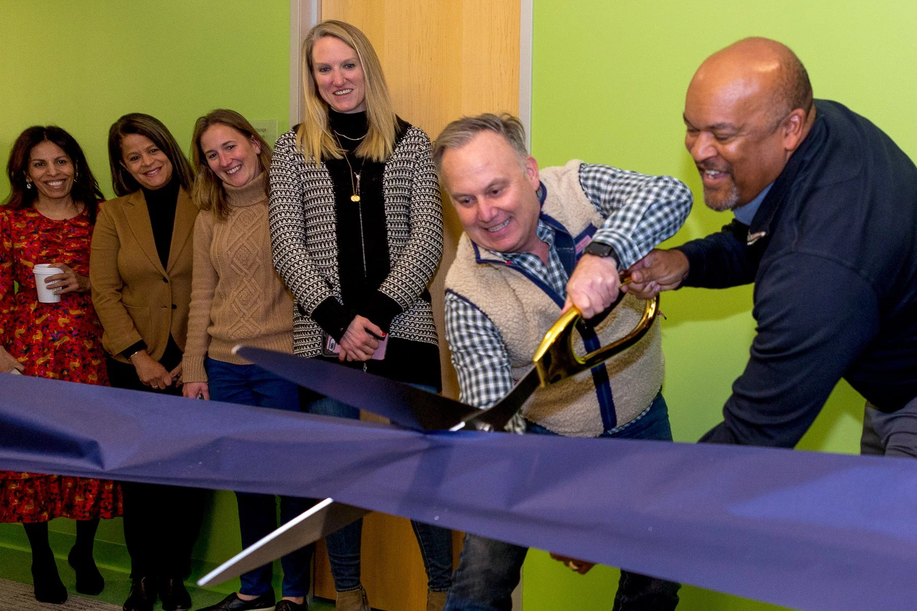 Group of six people gathered around a table, smiling, with one person cutting a ribbon.