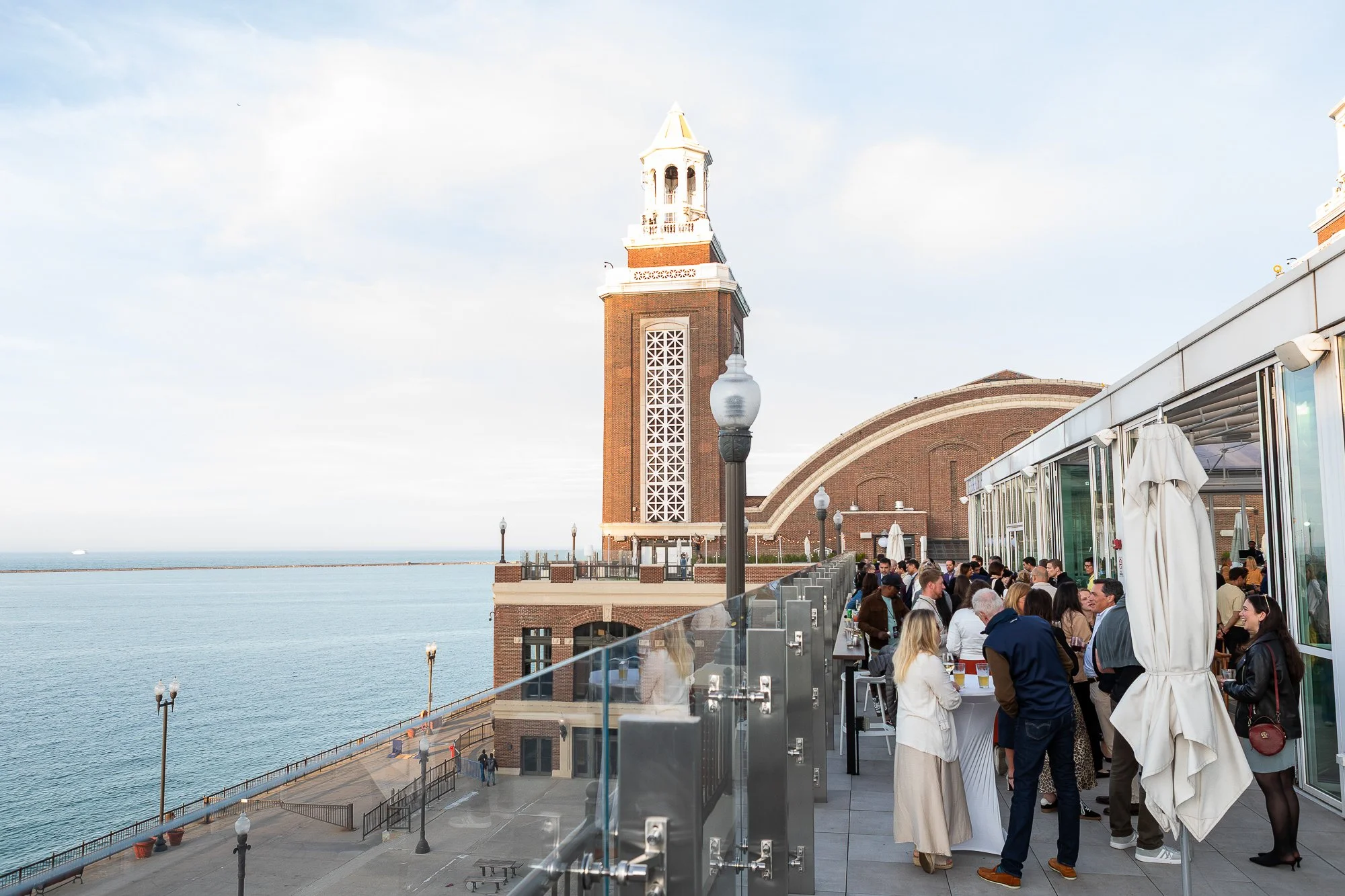 People gathered on an outdoor terrace overlooking the water, near a tall brick clock tower with a white top and decorative windows, possibly at a waterfront dining or event space in the late afternoon.©2026 Steven Barston