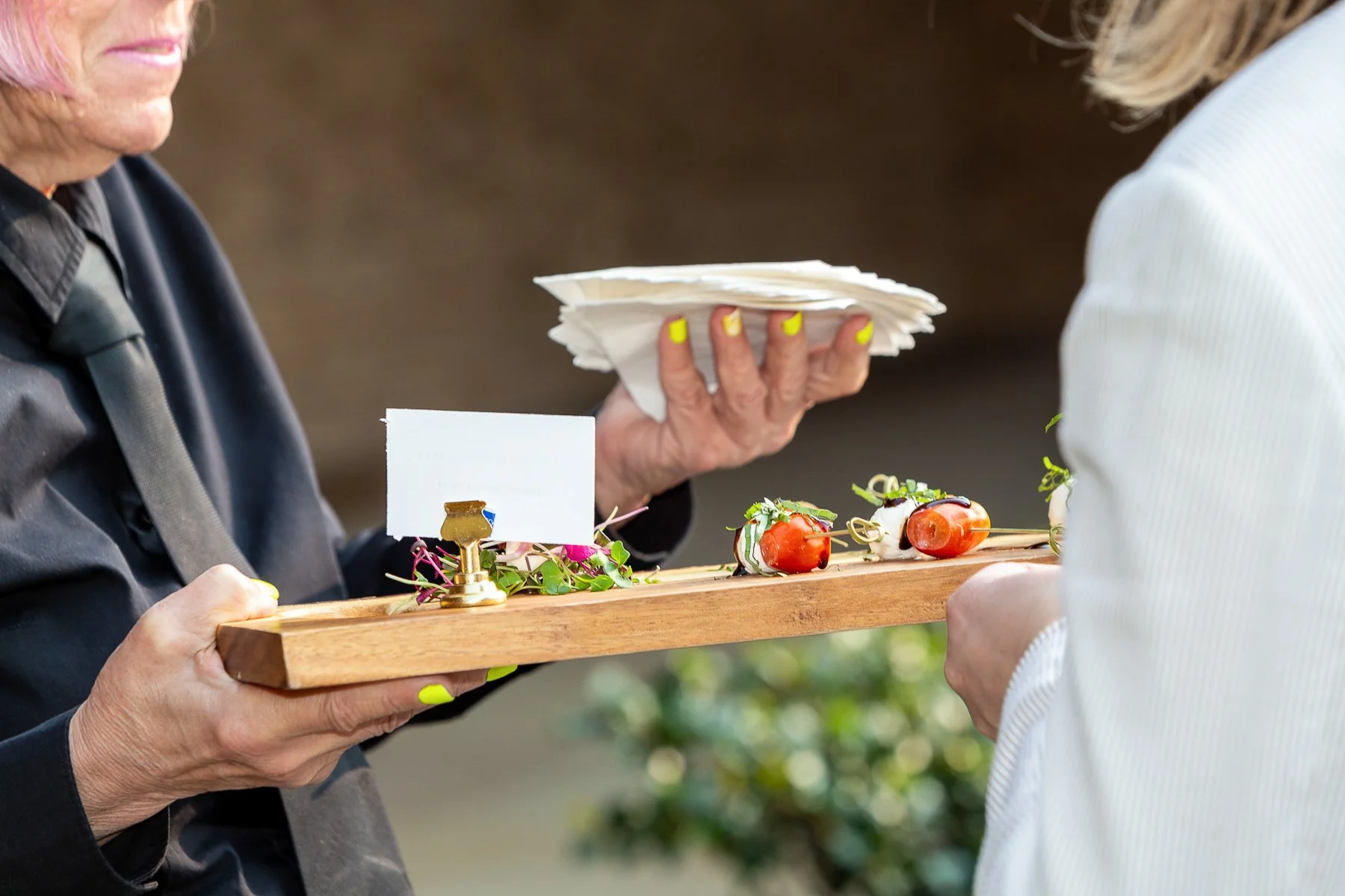 Two women exchanging a wooden tray of appetizers, including skewered cherry tomatoes and cheese, at an outdoor event.