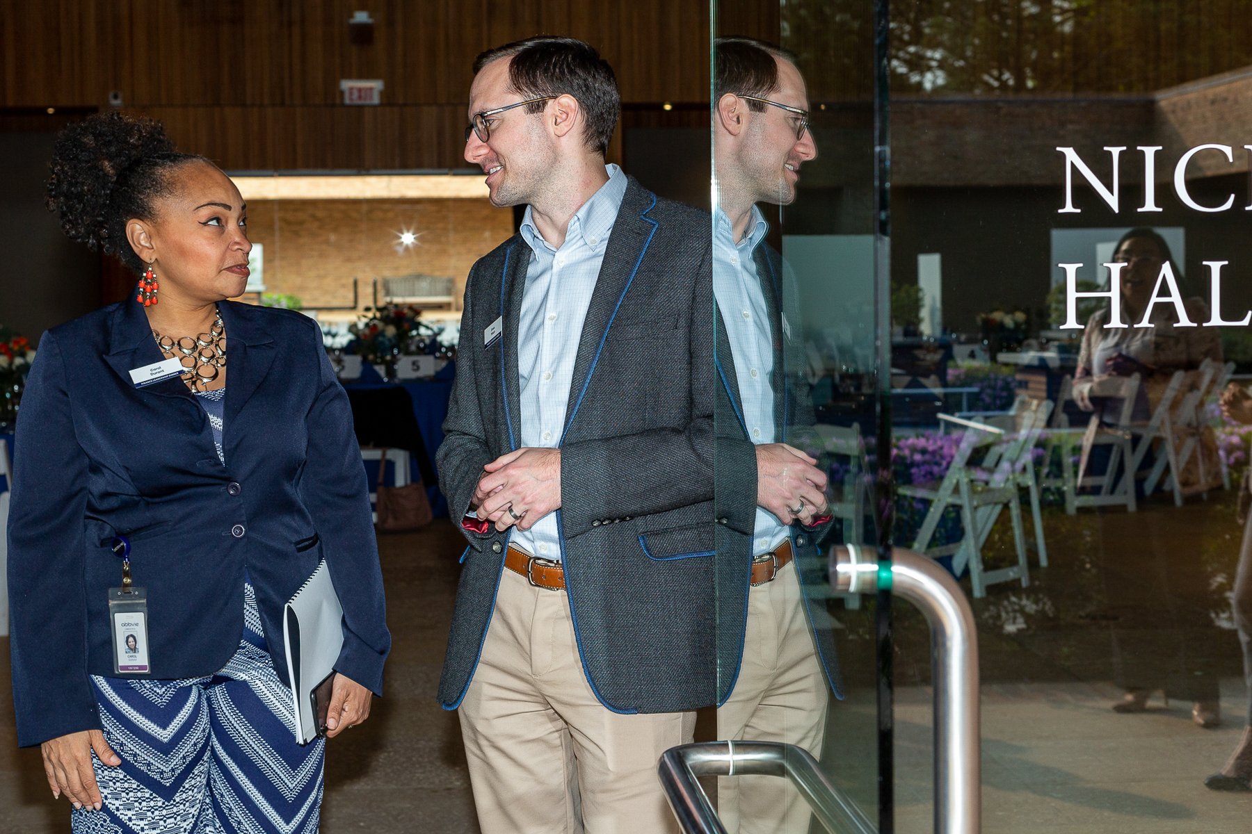 Two professionals, a woman and a man, are standing and talking outside a building with glass doors. The woman has curly black hair in a bun and is wearing a navy blazer, patterned dress, and jewelry. The man has glasses, short brown hair, and is wear