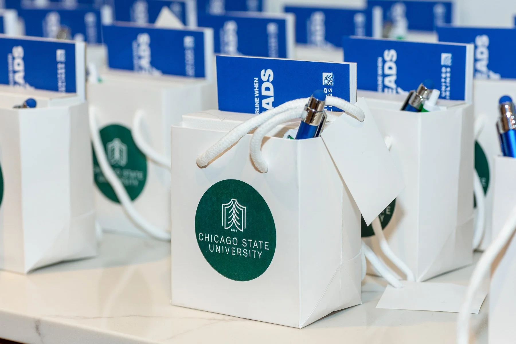 White gift bags with the Chicago State University logo arranged in rows, each containing pens and a blue educational flyer.