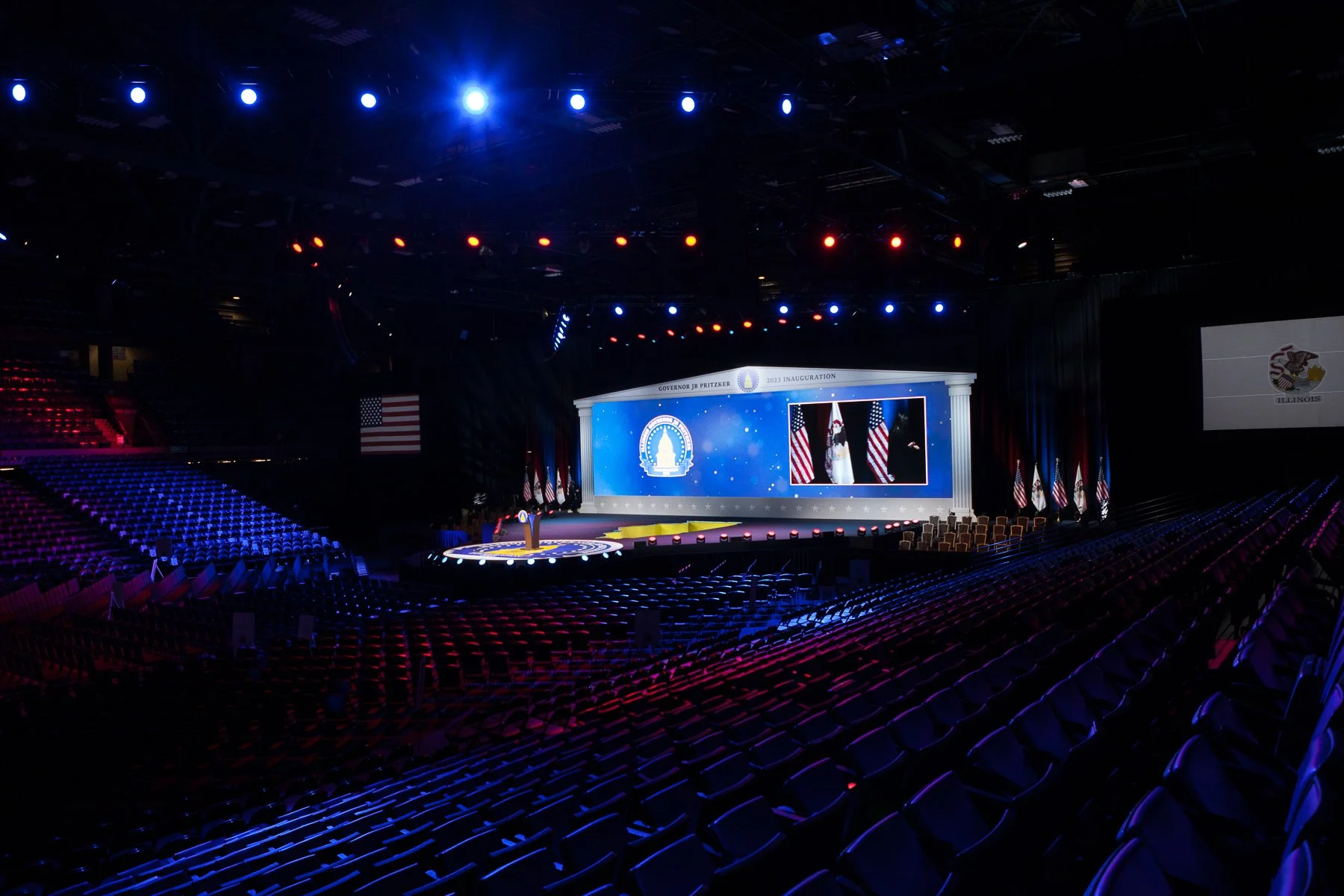 Large indoor hall prepared for a ceremony with a stage featuring a podium and large screens displaying official graphics, surrounded by American flags, with colorful lights illuminating the space and empty chairs arranged in rows for attendees.