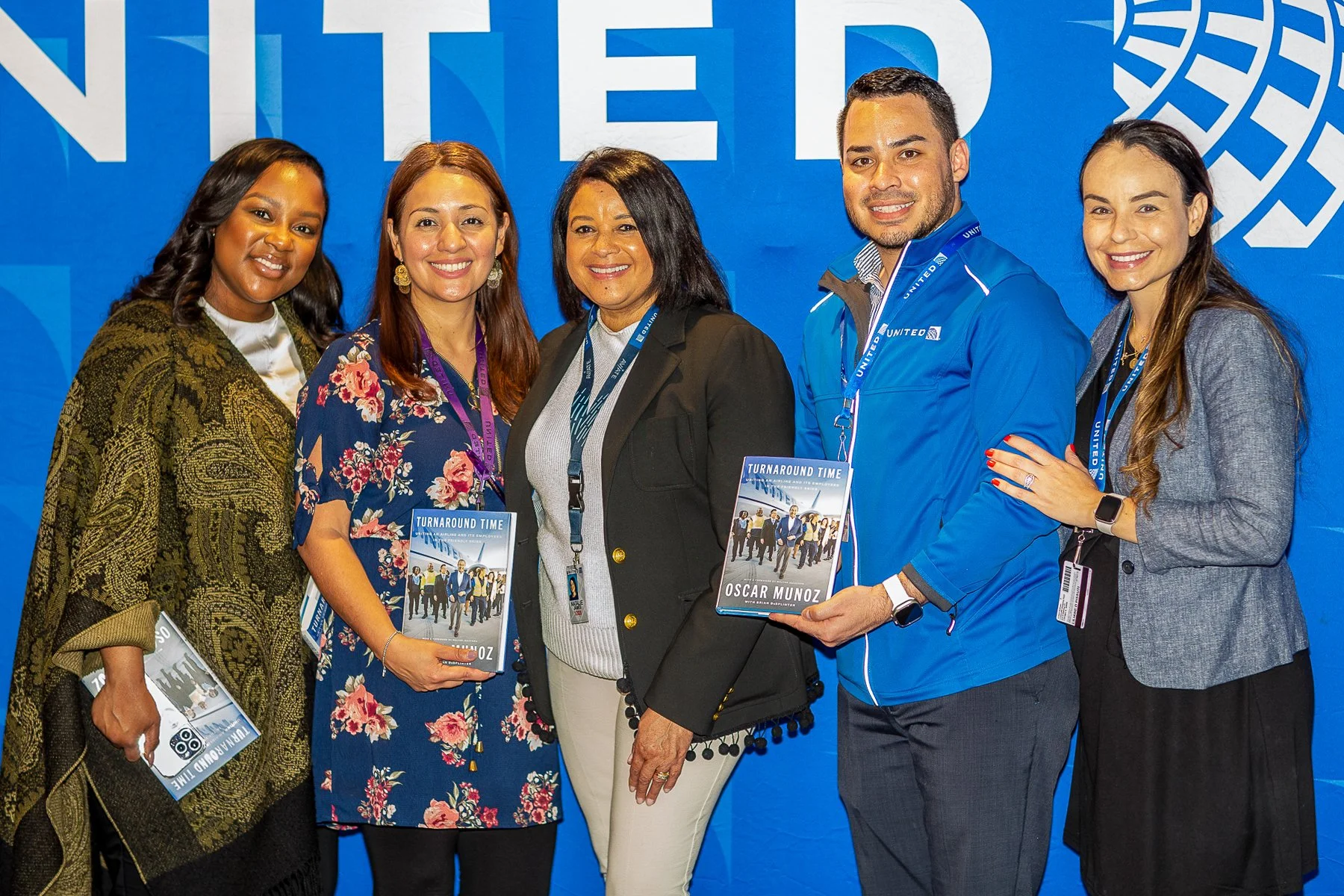 A group of six diverse people standing together at an event, smiling at the camera, with a blue backdrop behind them. One person is holding a book titled 'Turnaround Time' by Oscar Munoz.