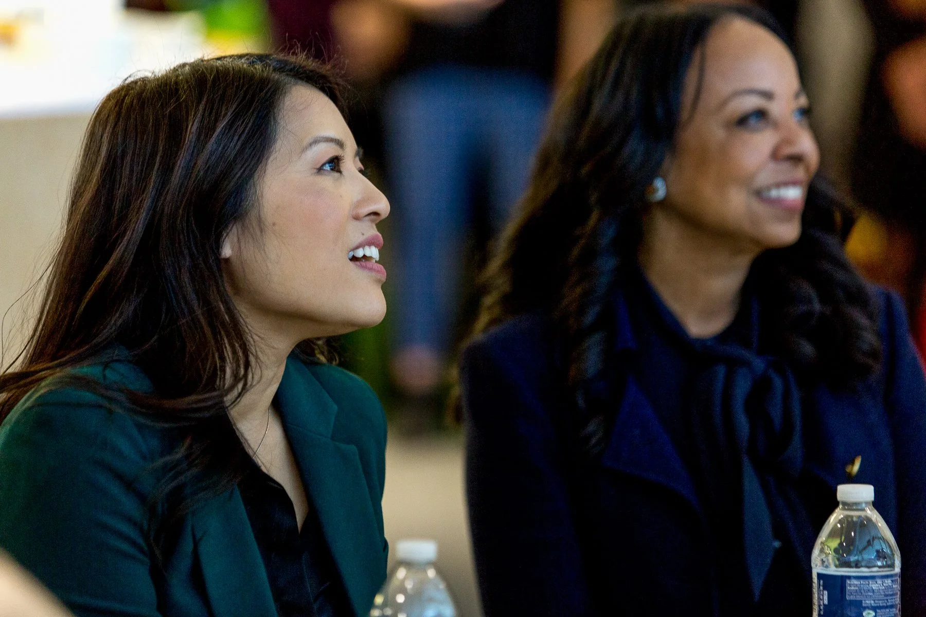 Two women sitting at a table, looking in the same direction and smiling. The woman on the left has long dark hair and is wearing a green blazer, while the woman on the right has long dark hair and is wearing a dark blue blazer.