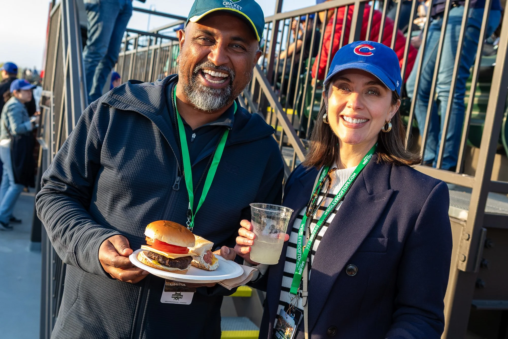 Two smiling people at an outdoor event, one man holding a plate with a burger and the woman holding a clear plastic cup, both wearing baseball caps and lanyards.©2026 Steven Barston