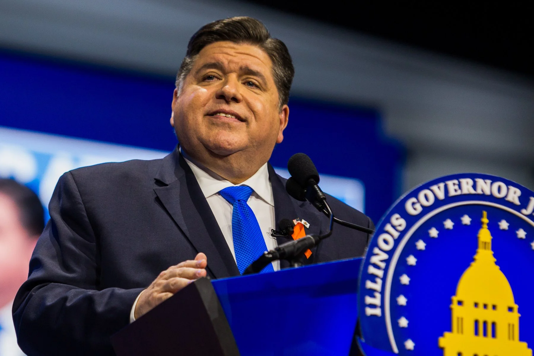 A man in a dark suit and blue tie speaking at a podium with a seal that reads 'Louisiana Governor' and featuring a building with a spire in the background.