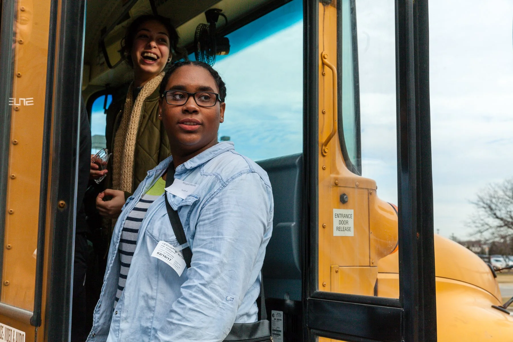 Two women leaving a school bus, one is looking away, and the other is smiling and looking off to the side, with trees and a cloudy sky in the background.