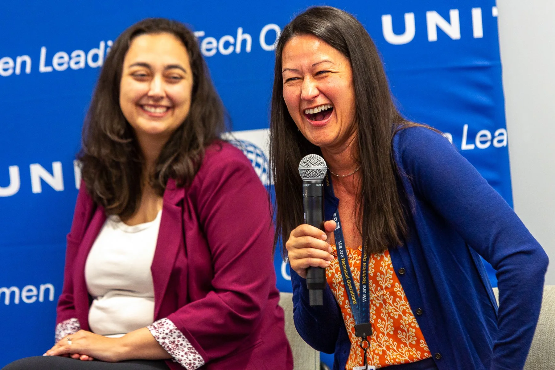 Two women laughing and smiling during a speaking event, one holding a microphone, in front of a blue backdrop with white text.©2026 Steven Barston