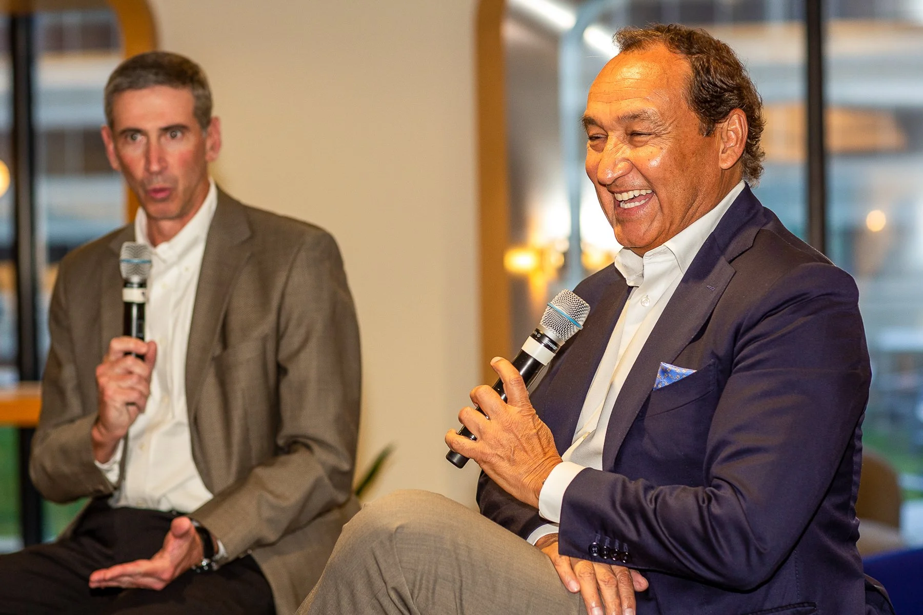 Two men in suits holding microphones during a discussion or interview, seated indoors with large windows in the background.